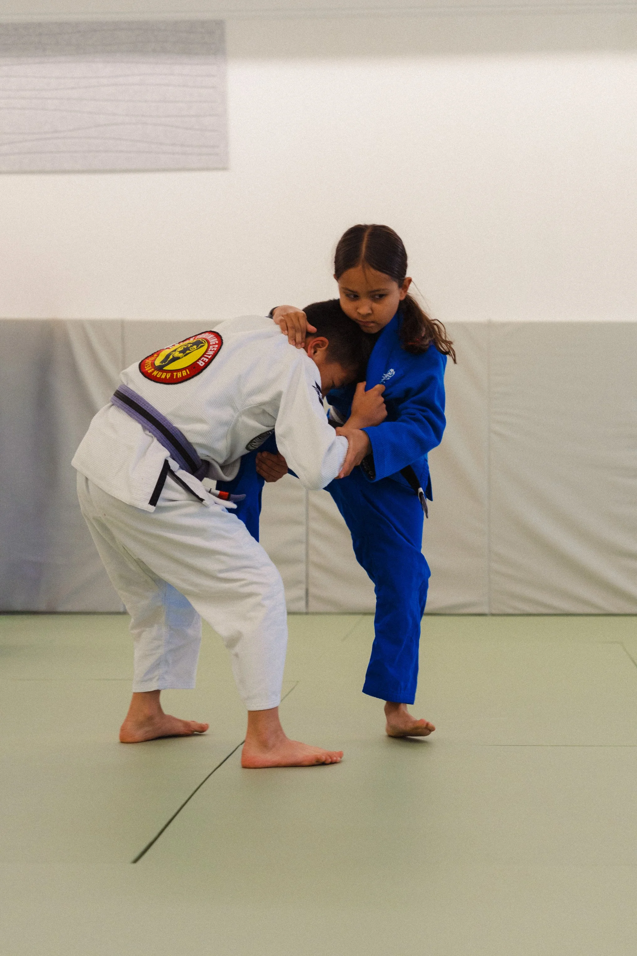 Two children practicing Brazilian Jiu-Jitsu in a dojo, wearing gis and focusing on a grappling move.