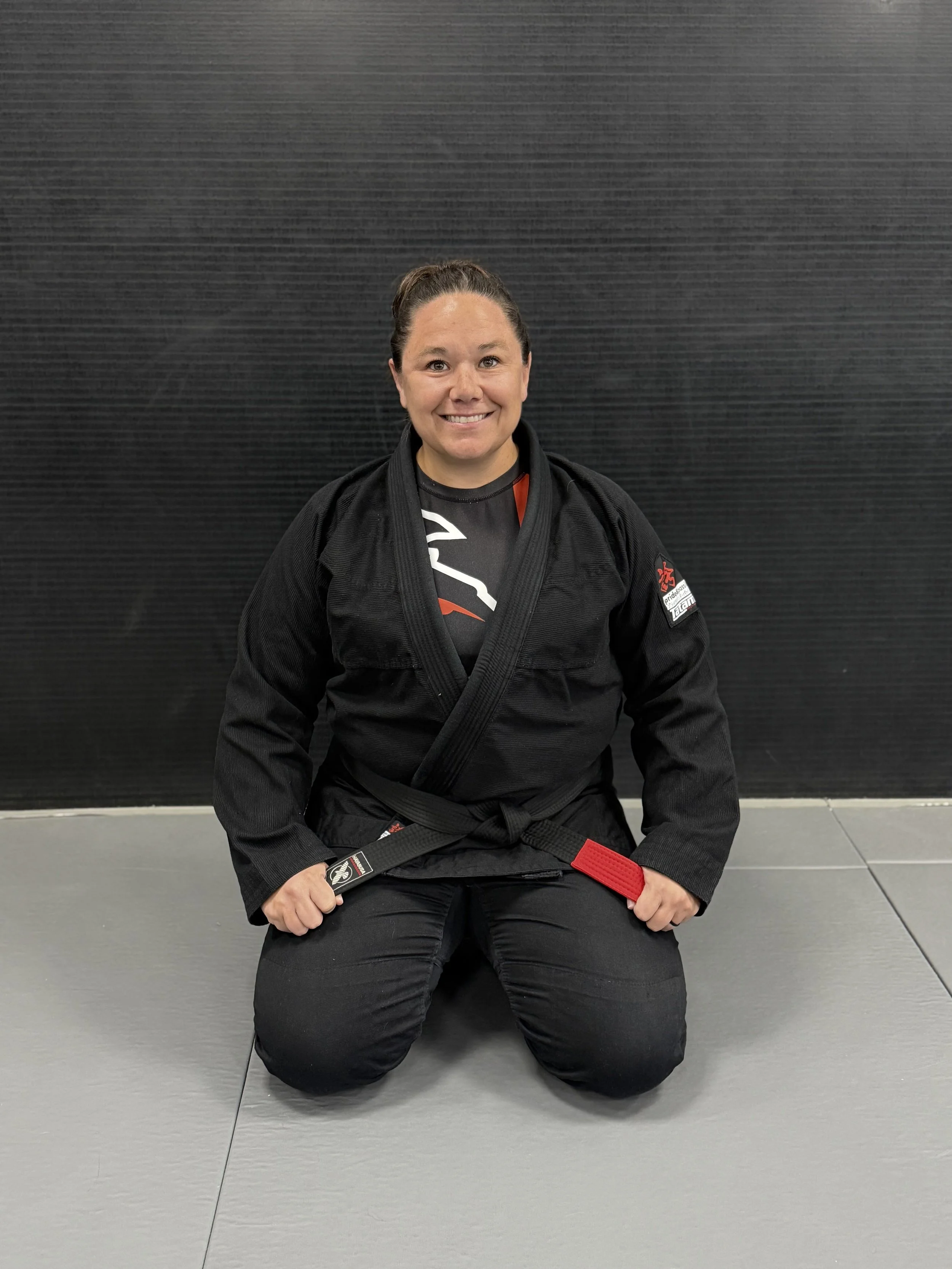 A woman in a black Brazilian Jiu-Jitsu gi kneeling on the mat with a smile, holding her black belt with a red tab, in front of a black wall.