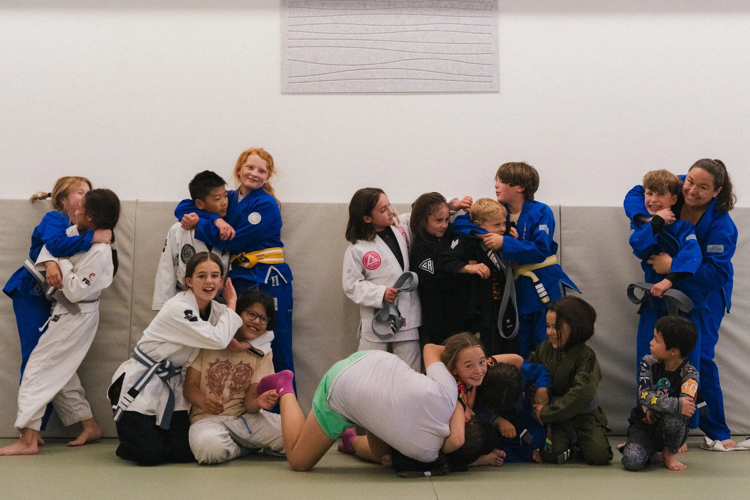 Group of children and a woman in judo uniforms standing and sitting on a mat in a sports hall, smiling and hugging each other after a judo class.