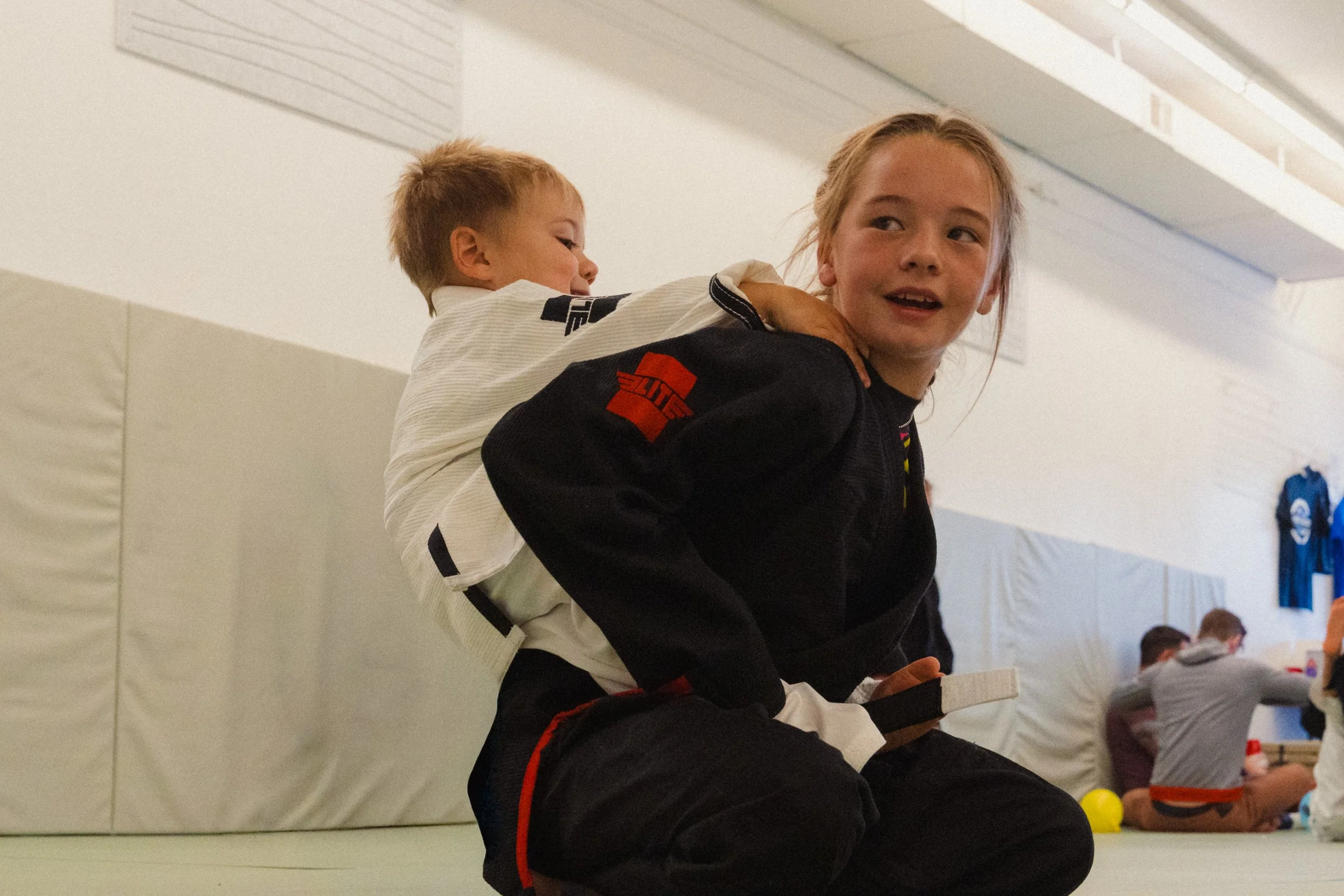 A young girl is sitting cross-legged on the floor in a martial arts uniform, smiling and looking to her right, with a young boy on her back in a martial arts gi, smiling and looking to her left, inside a martial arts training facility.