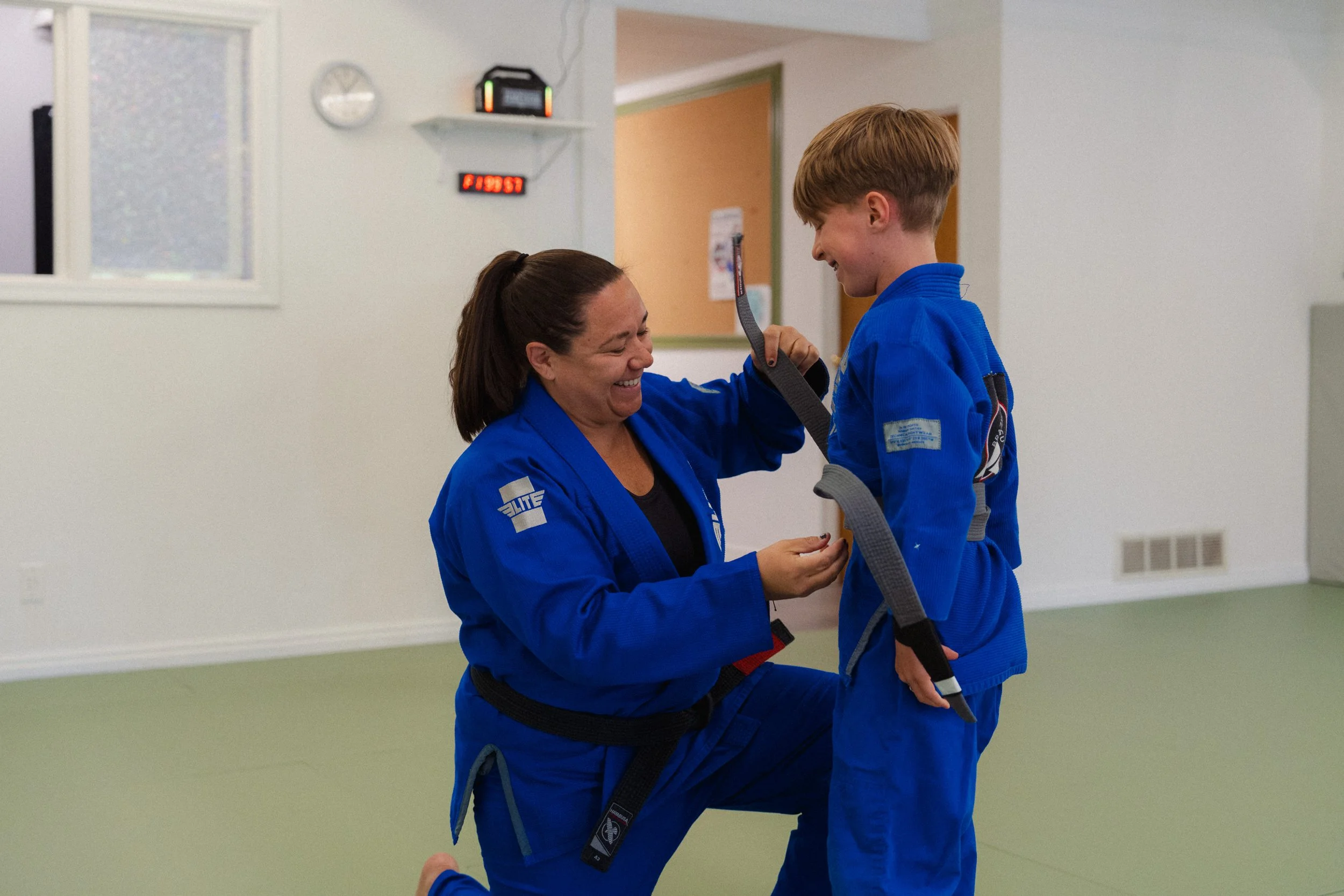 A woman in a blue martial arts uniform is smiling and holding the strap of a young boy's martial arts uniform, who is also dressed in blue.