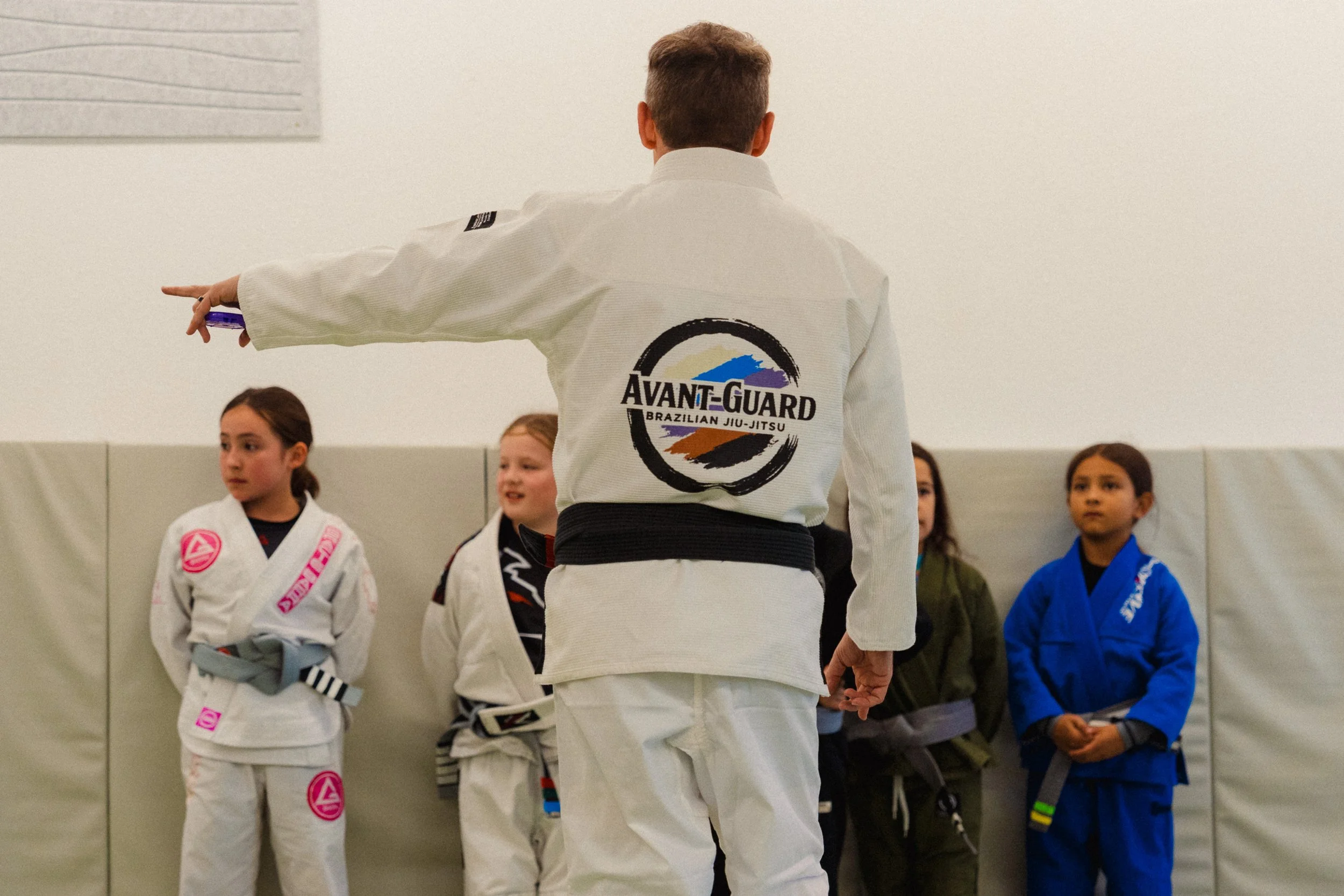 A martial arts instructor in a white gi with an 'AVANT-GUARD' logo on the back demonstrating a technique to young students in gi uniforms, standing against a plain wall with other children watching.