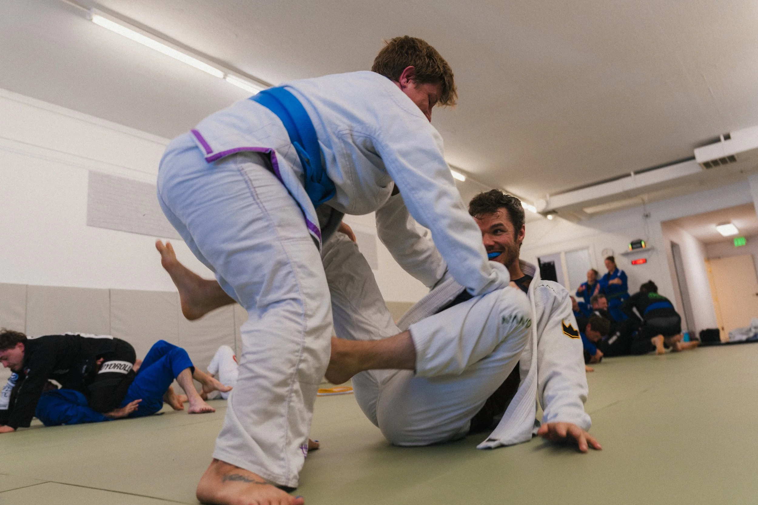 Two martial artists practicing Brazilian Jiu-Jitsu on a padded mat in a training facility. One person is executing a grappling move on another, who is lying on the floor, with several other practitioners visible in the background.