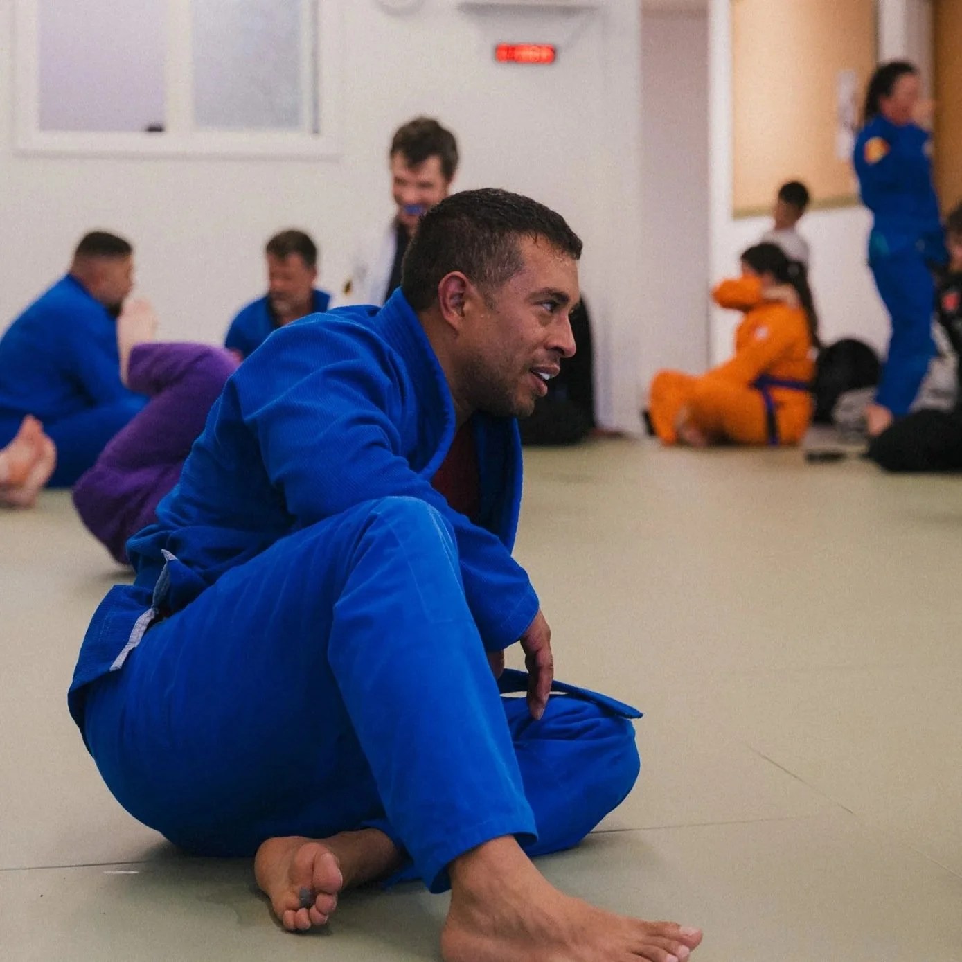 A man in a blue BJJ gi sitting on the mat with his right leg bent in front of him and his left leg extended, in Avant-Guard Brazillian Jiu Jitsu with other practitioners in gi and a person in an orange gi seated against the wall in the background.