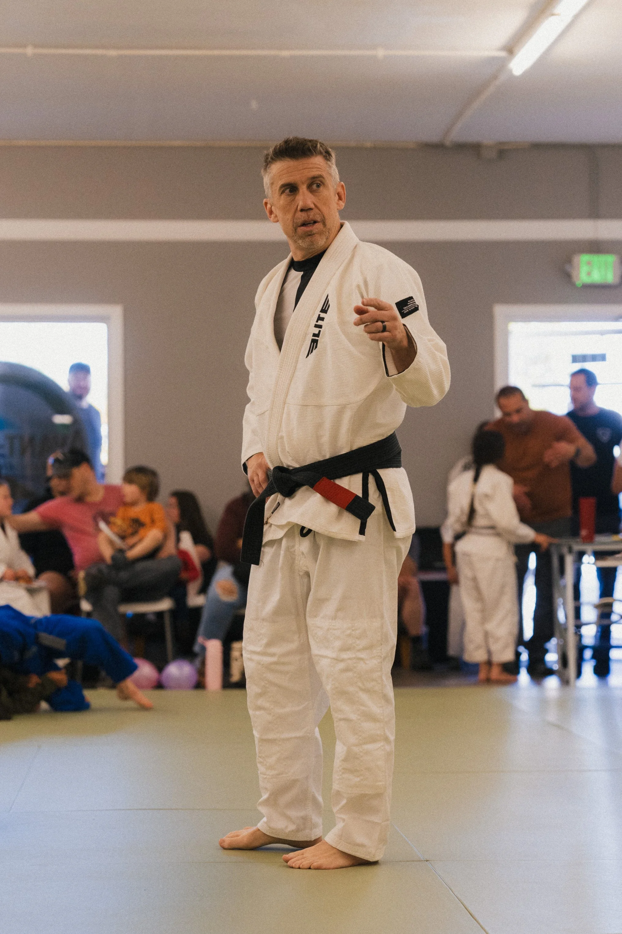 Man in a martial arts gi with a black belt demonstrating a technique at a martial arts class, indoors, with students seated in the background.