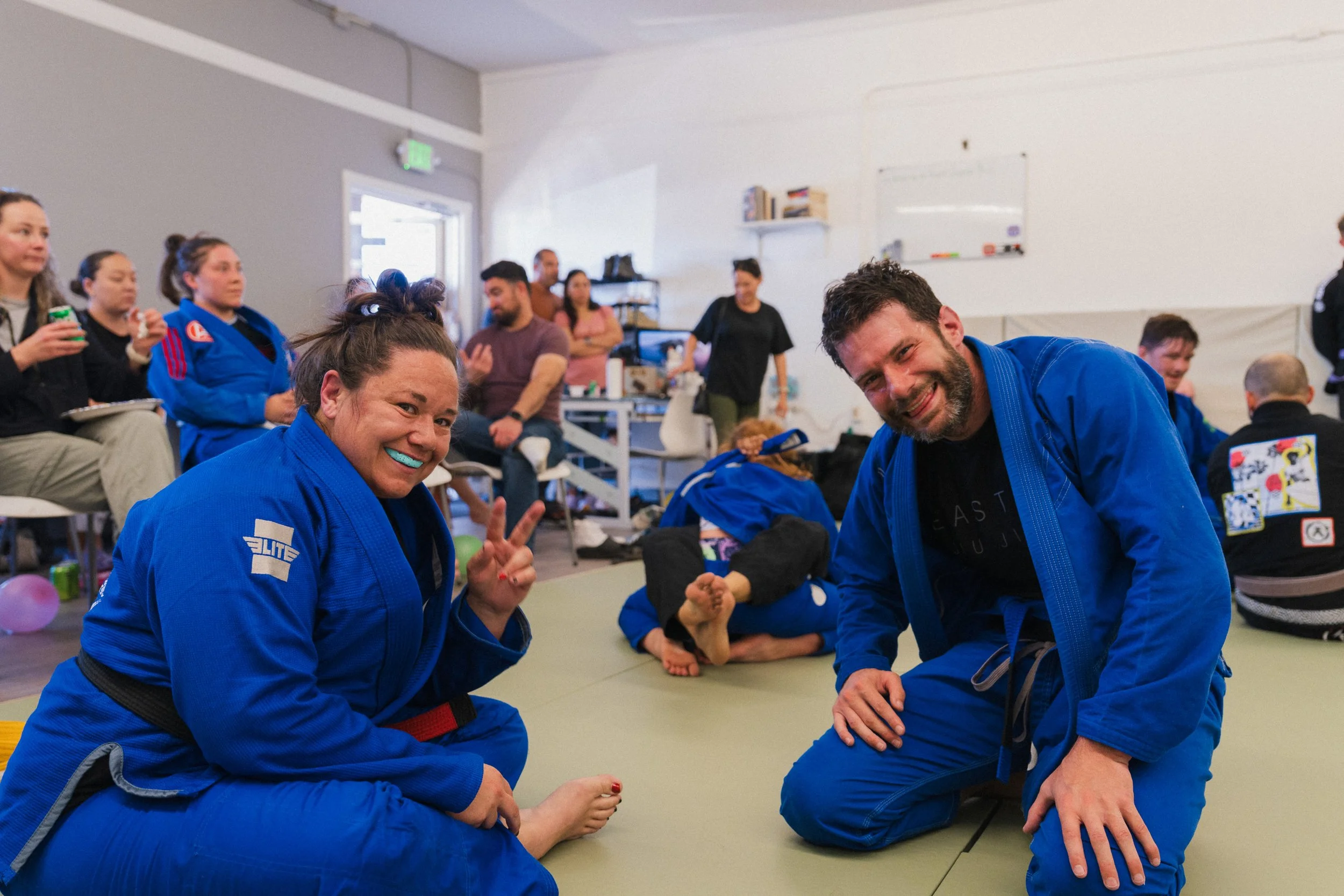 A group of martial arts students in blue gis, sitting and kneeling on a mat in a dojo, with some smiling and making peace signs. In the background, other students and an instructor are visible, with some seated and others standing, in a well-lit room