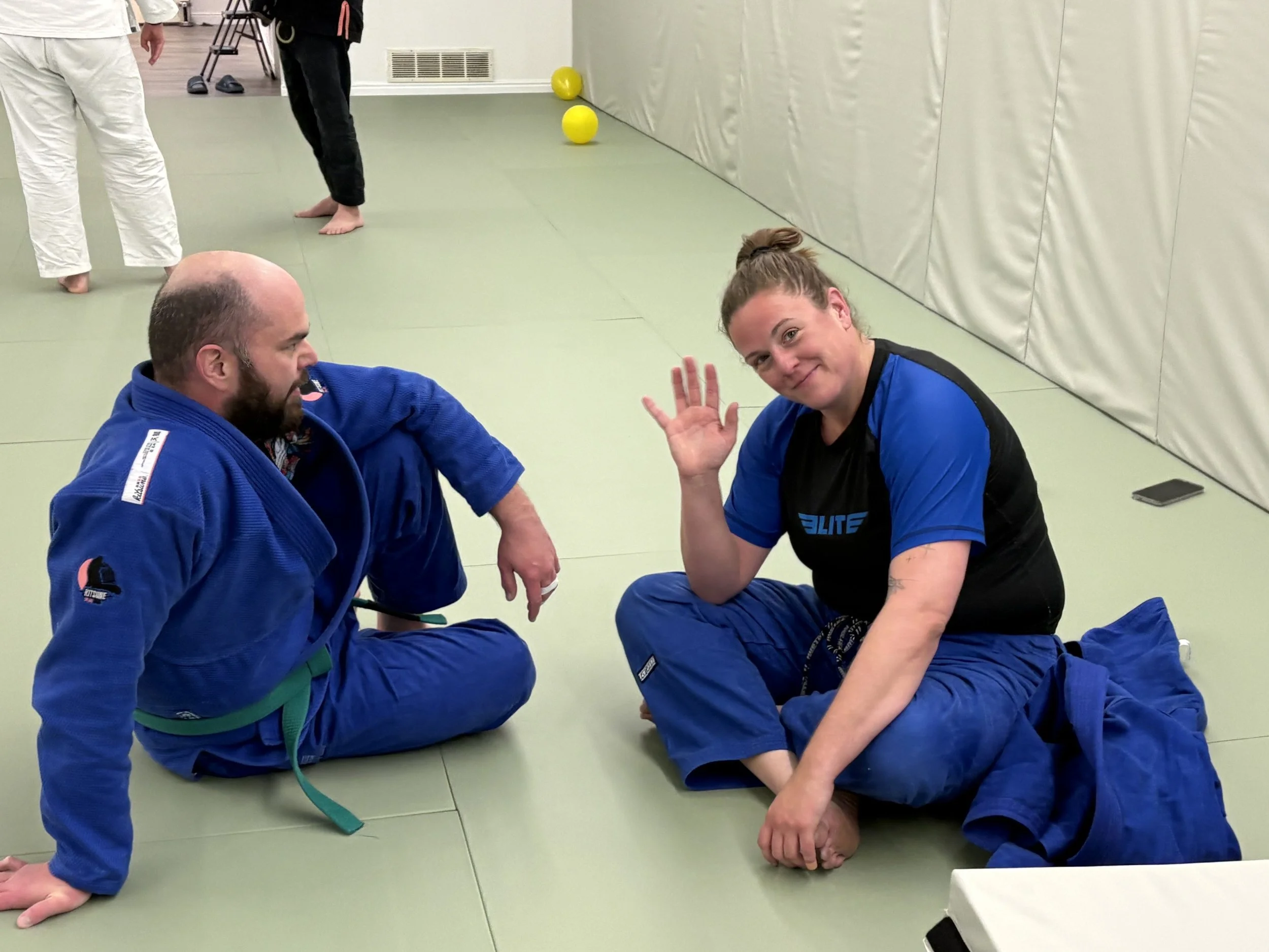 Two Brazilian Jiu-Jitsu practitioners, one man and one woman, practicing on a mat; the woman is waving at the camera and both are dressed in gi uniforms.