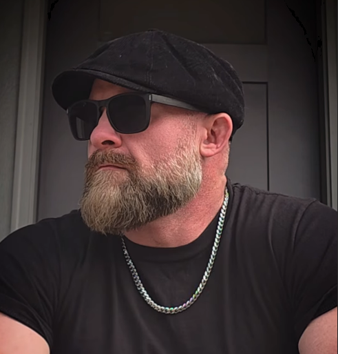 A man with a beard wearing sunglasses, a black cap, a black T-shirt, and a silver chain necklace, sitting outdoors.