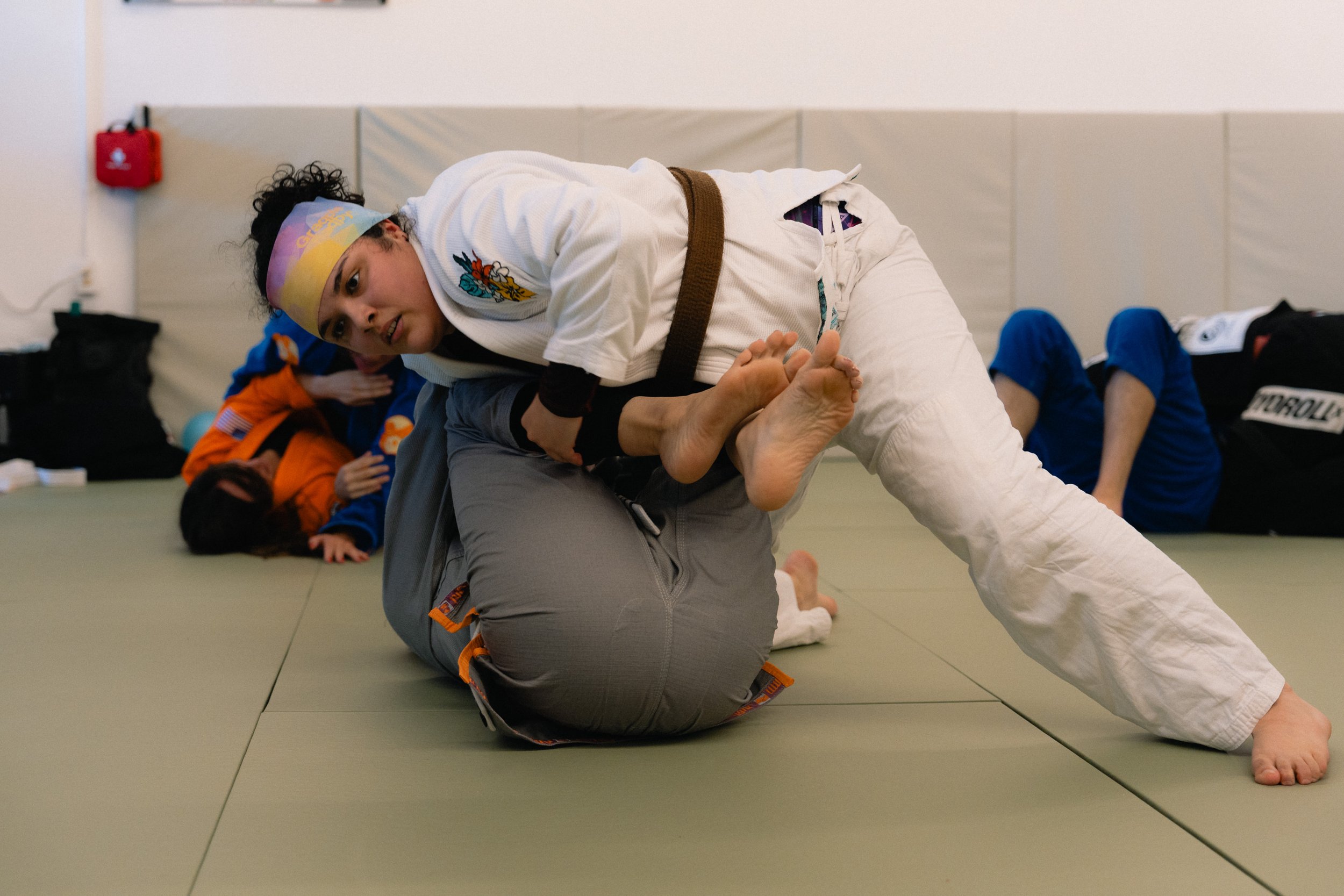 Two people practicing Brazilian Jiu-Jitsu on a green mat indoors. One person, in a white gi with a brown belt, is executing a ground technique on another person in a gray martial arts uniform. Other participants in gis are visible in the background.