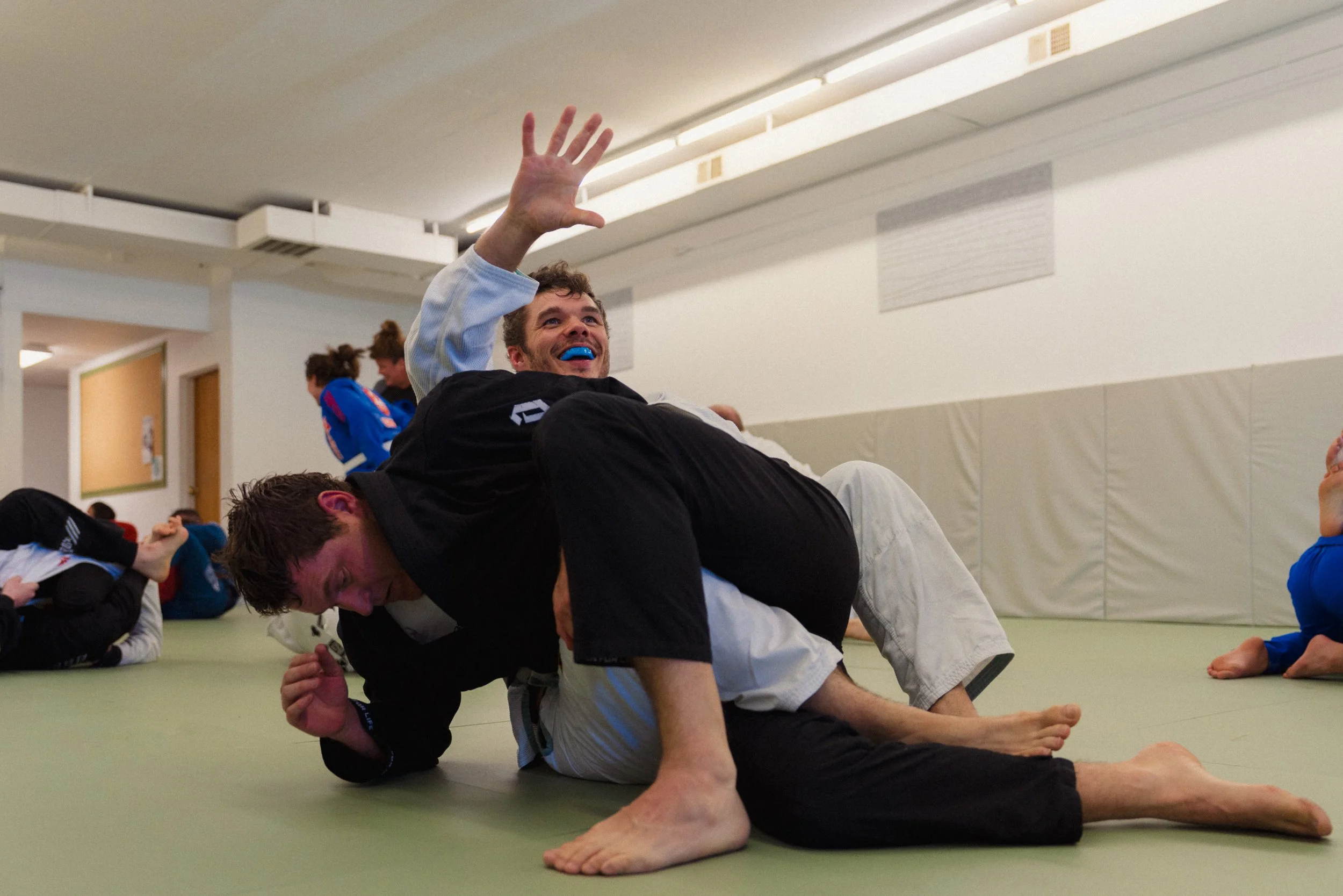 People practicing Brazilian Jiu-Jitsu on a mat, with two participants in the foreground engaged in a grappling move, one on top of the other, in a large indoor gym or dojo.