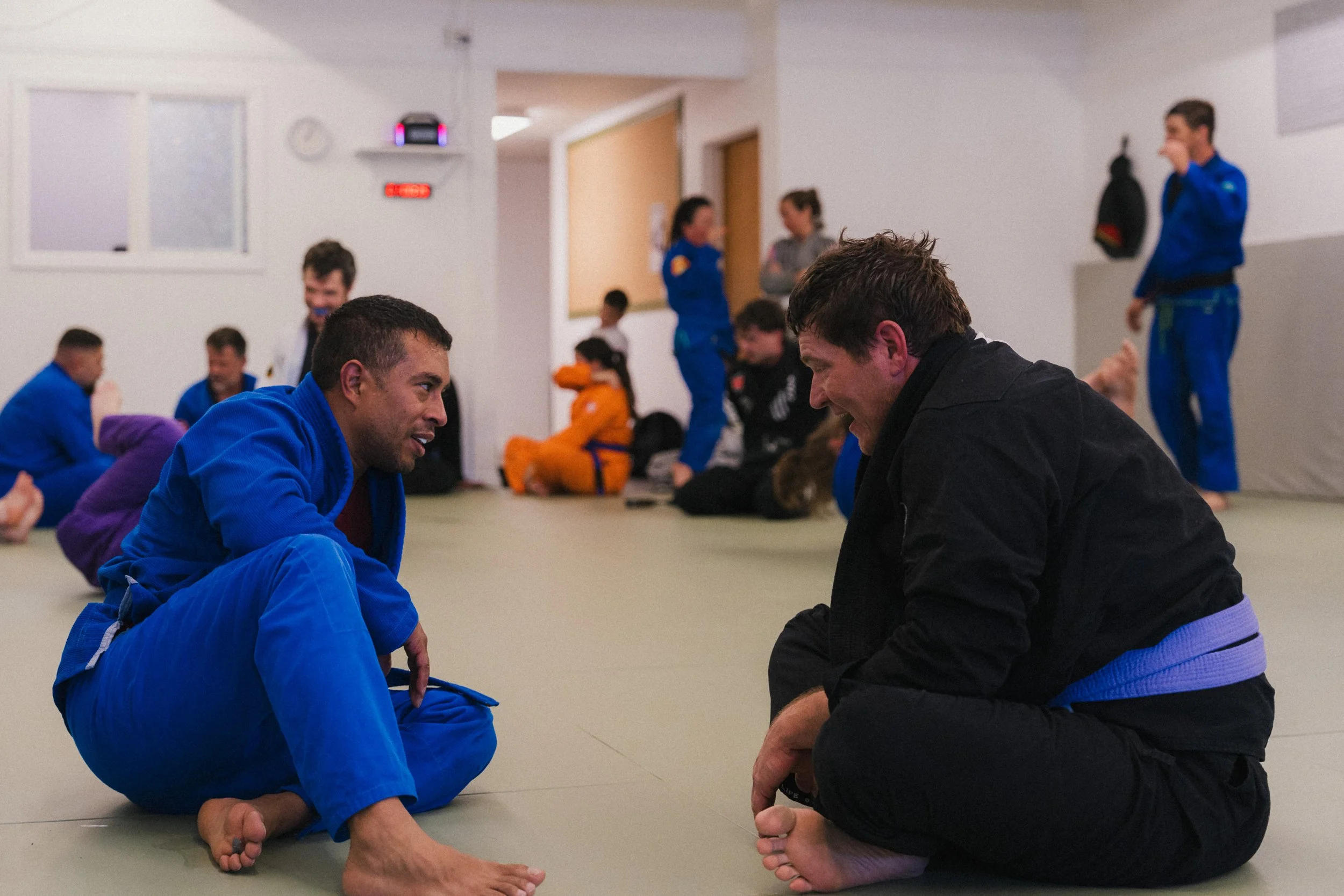 Two men dressed in martial arts uniforms engage in a friendly conversation while sitting cross-legged on a mat in a martial arts training facility, with other students and instructors in the background.