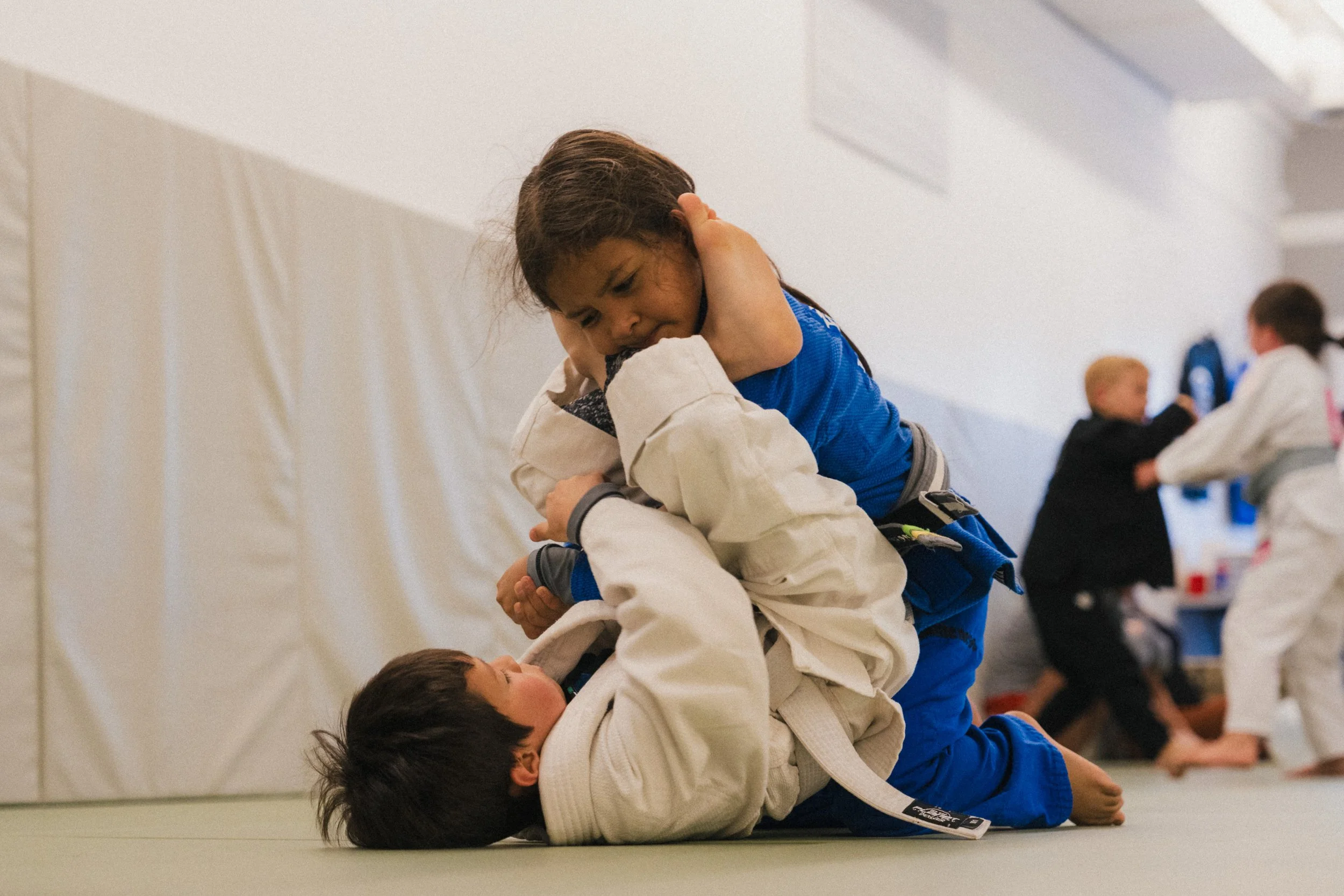 Two children practicing Brazilian Jiu-Jitsu on mats, with one child in a blue gi on top of the child in a white gi, who is lying on the ground.