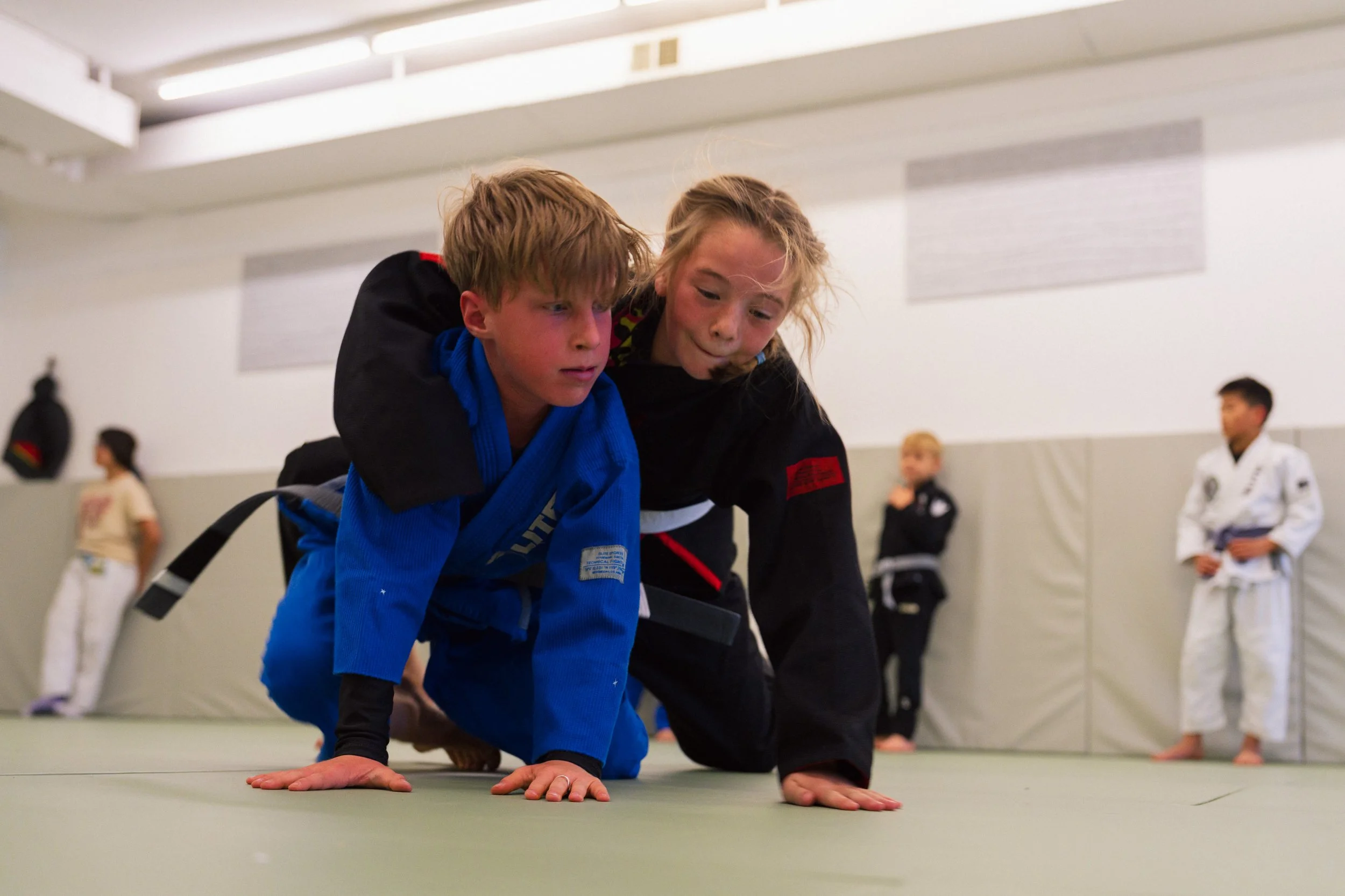 Two children practicing Brazilian Jiu-Jitsu on the training mat in a martial arts gym, with other students and a punching bag visible in the background.