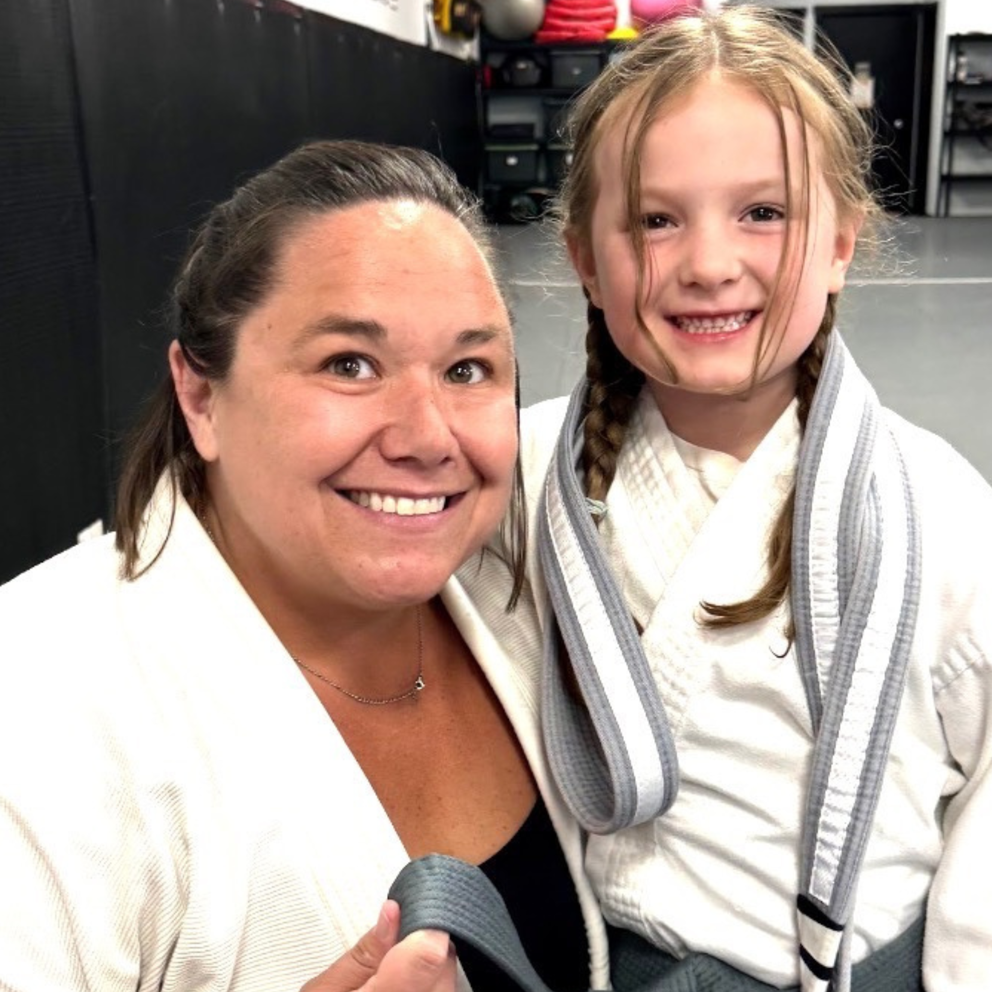 A woman in a white shirt and a girl in a gray hoodie with braids smiling for a photo at a gym, with a hand holding a black massage roller in the foreground.