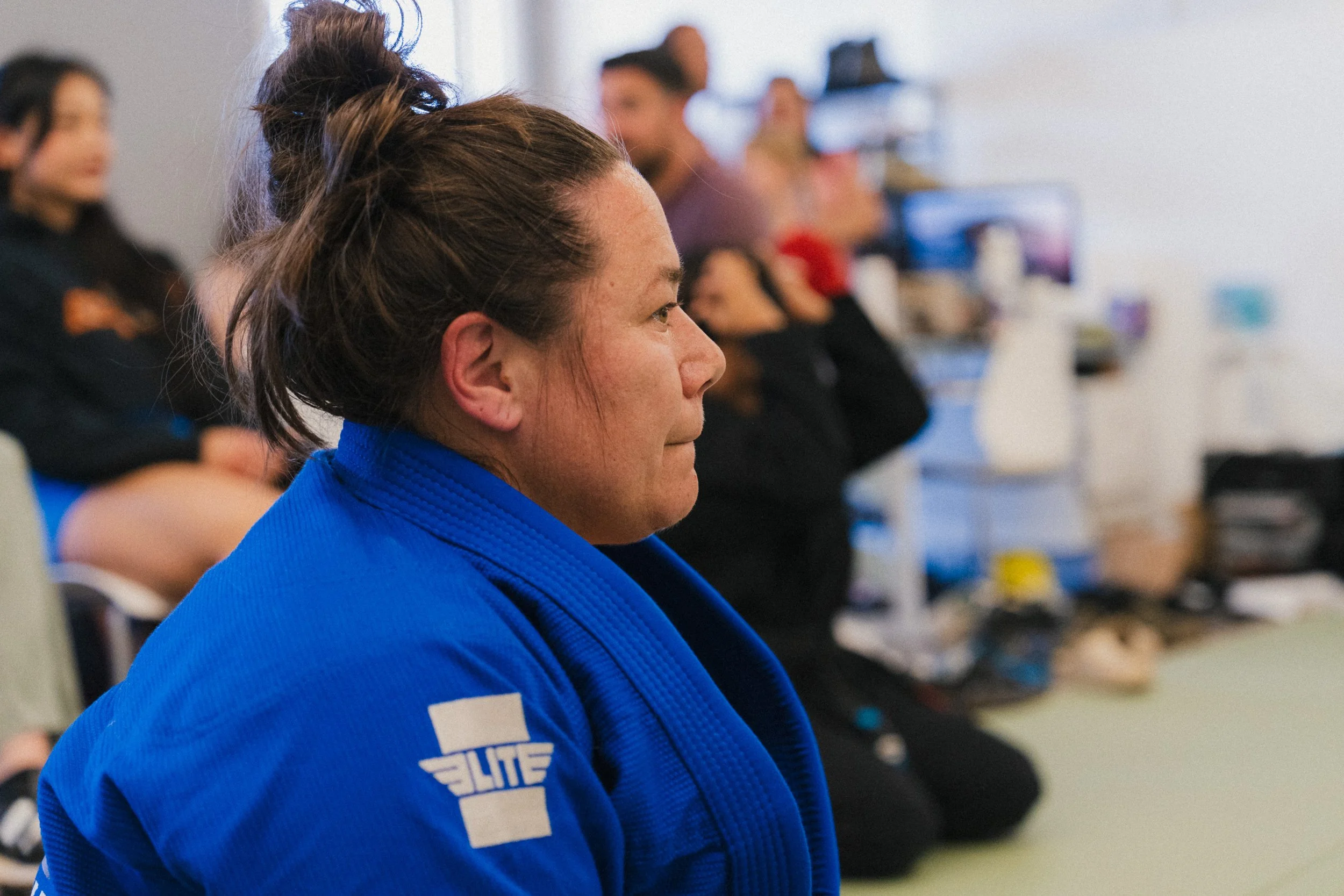A woman with brown hair in a bun wearing a blue martial arts gi, sitting in a room with other women also dressed in similar attire, likely in a martial arts class or seminar.