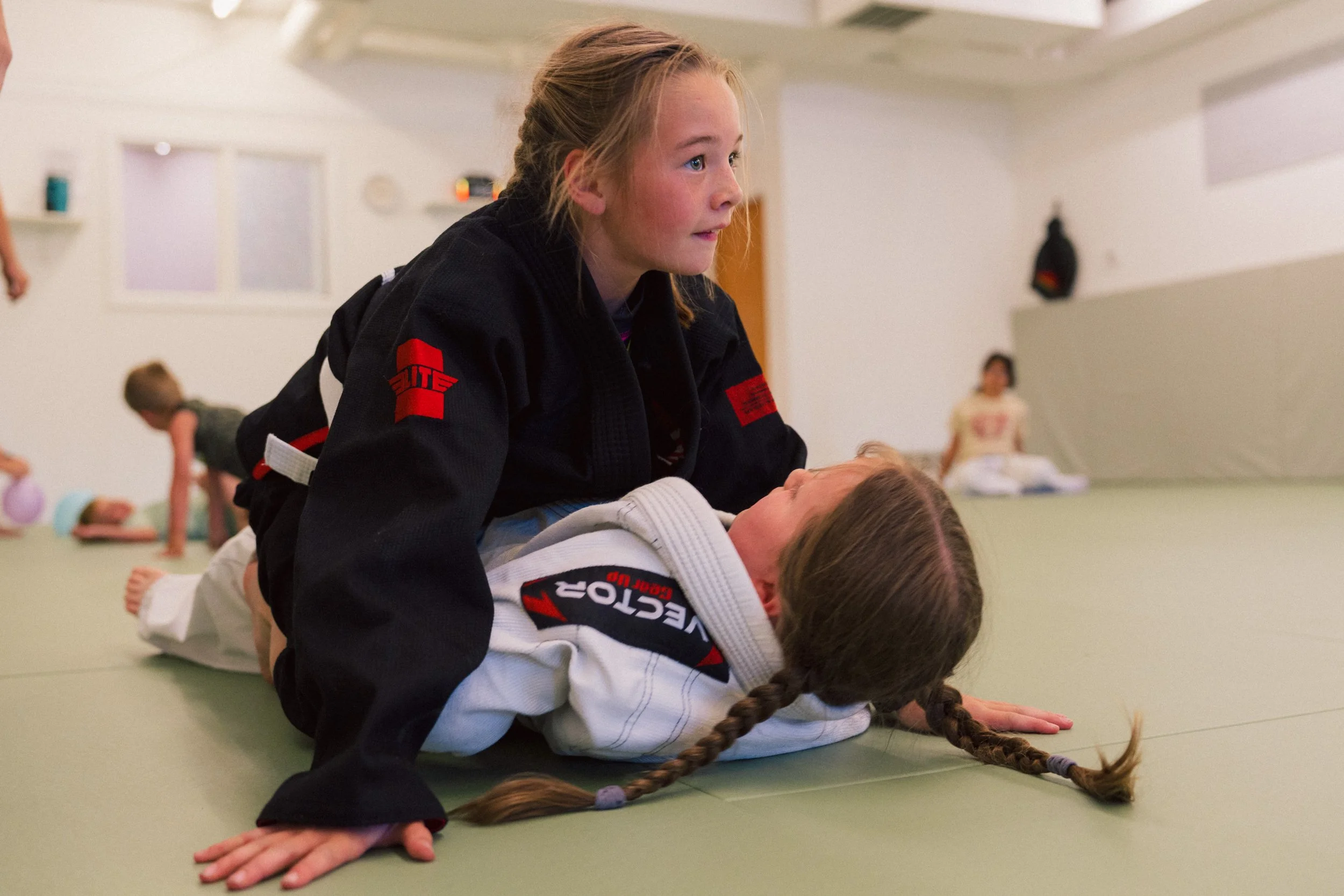 A girl in a martial arts gi with a red patch on the sleeve instructs a young girl in a white gi during a Brazilian Jiu-Jitsu class; they are on a green mat in a gym with children practicing in the background.