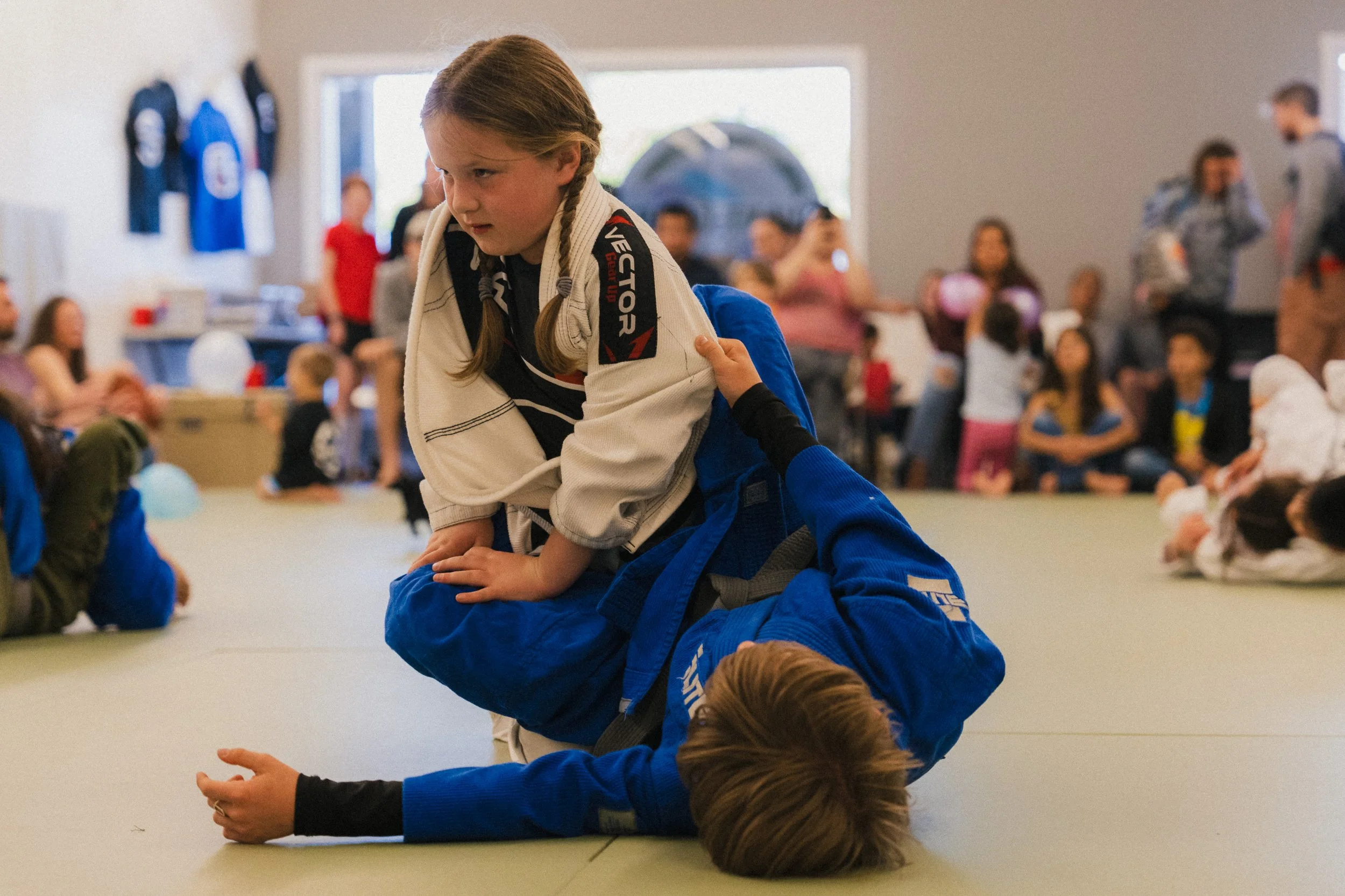 A young girl practicing Brazilian Jiu-Jitsu in a gym, wearing a white gi, during a training session or class, with a crowd of people and other children in the background.