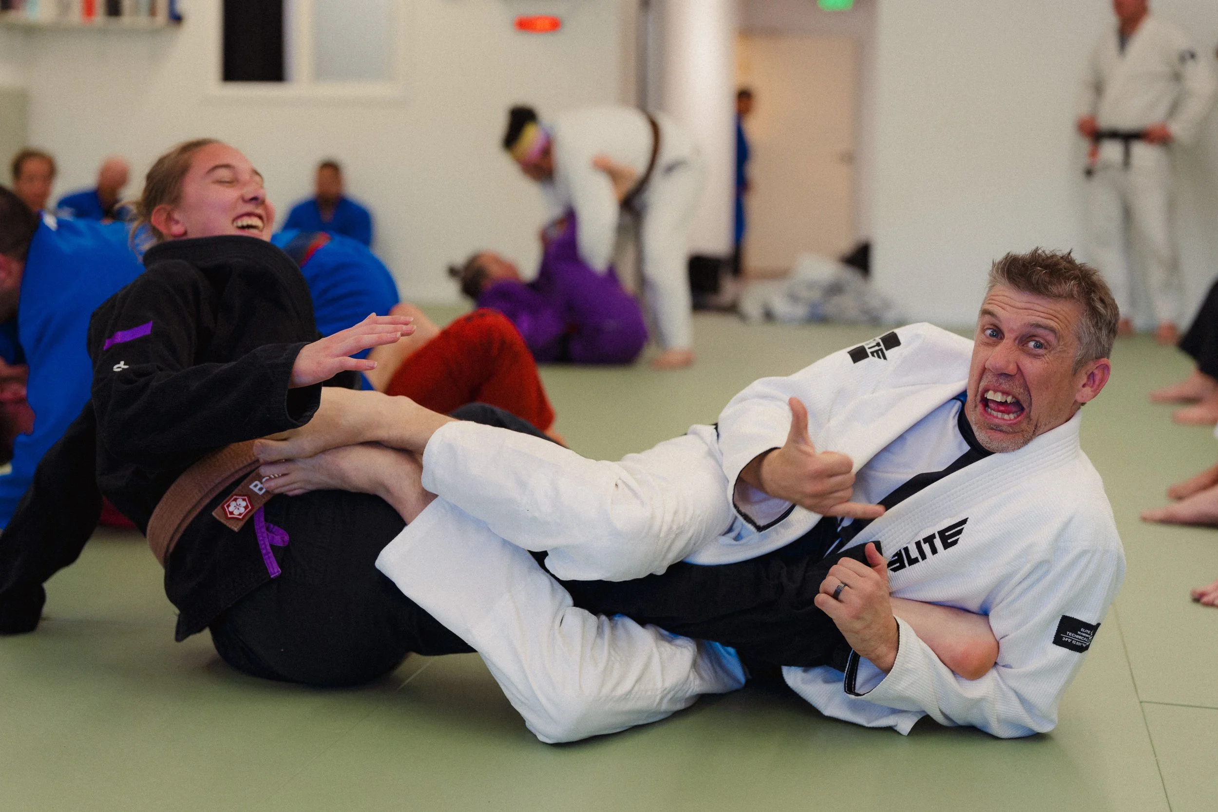 Two martial artists practicing Brazilian Jiu-Jitsu on a green mat, with one in a black gi and the other in a white gi, smiling and giving a thumbs-up during training or competition in a gym surrounded by other students.