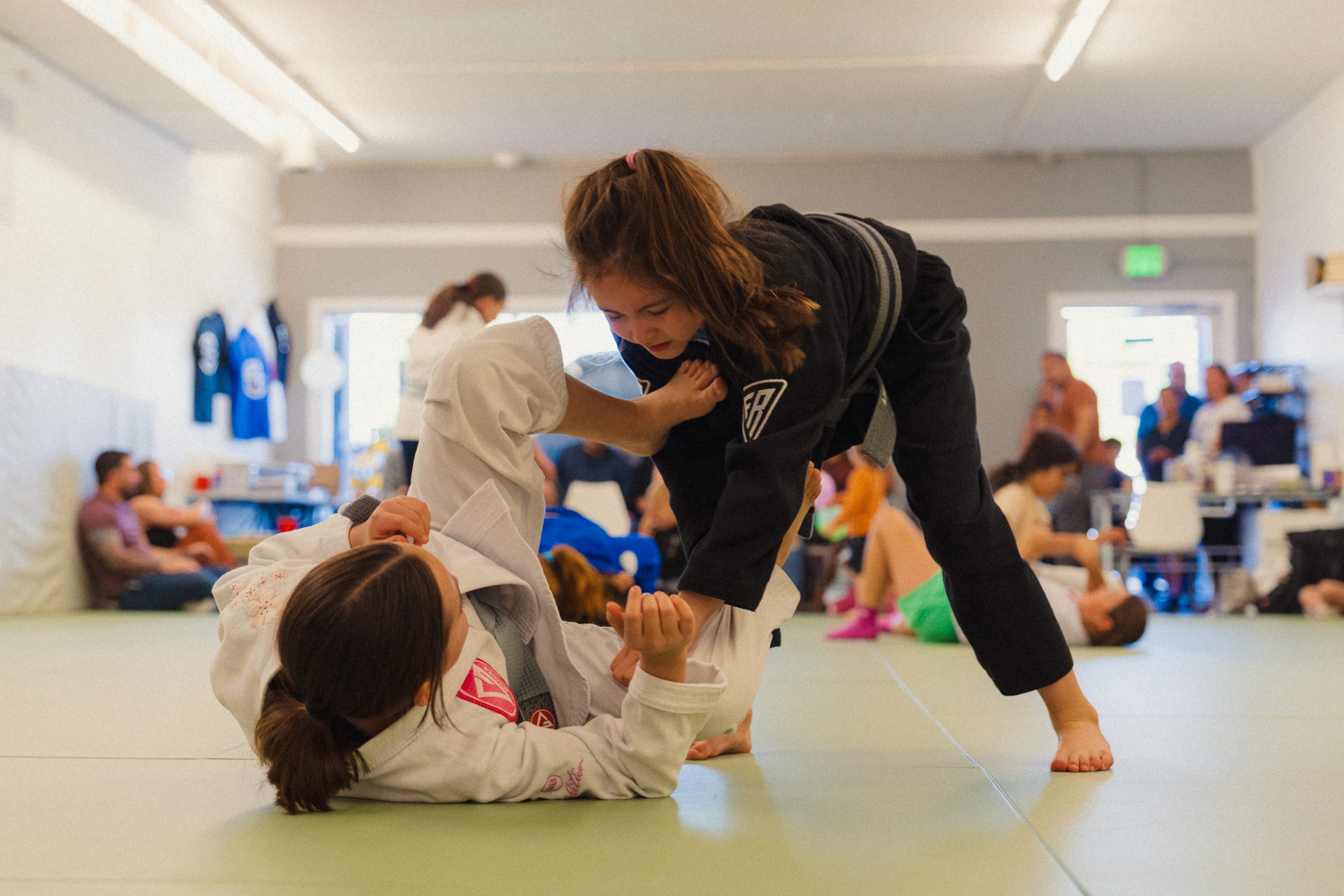 Two young girls practicing Brazilian Jiu-Jitsu, one girl in a white gi on the ground and the other in a black gi on top, during a class in a dojo with other students in the background.