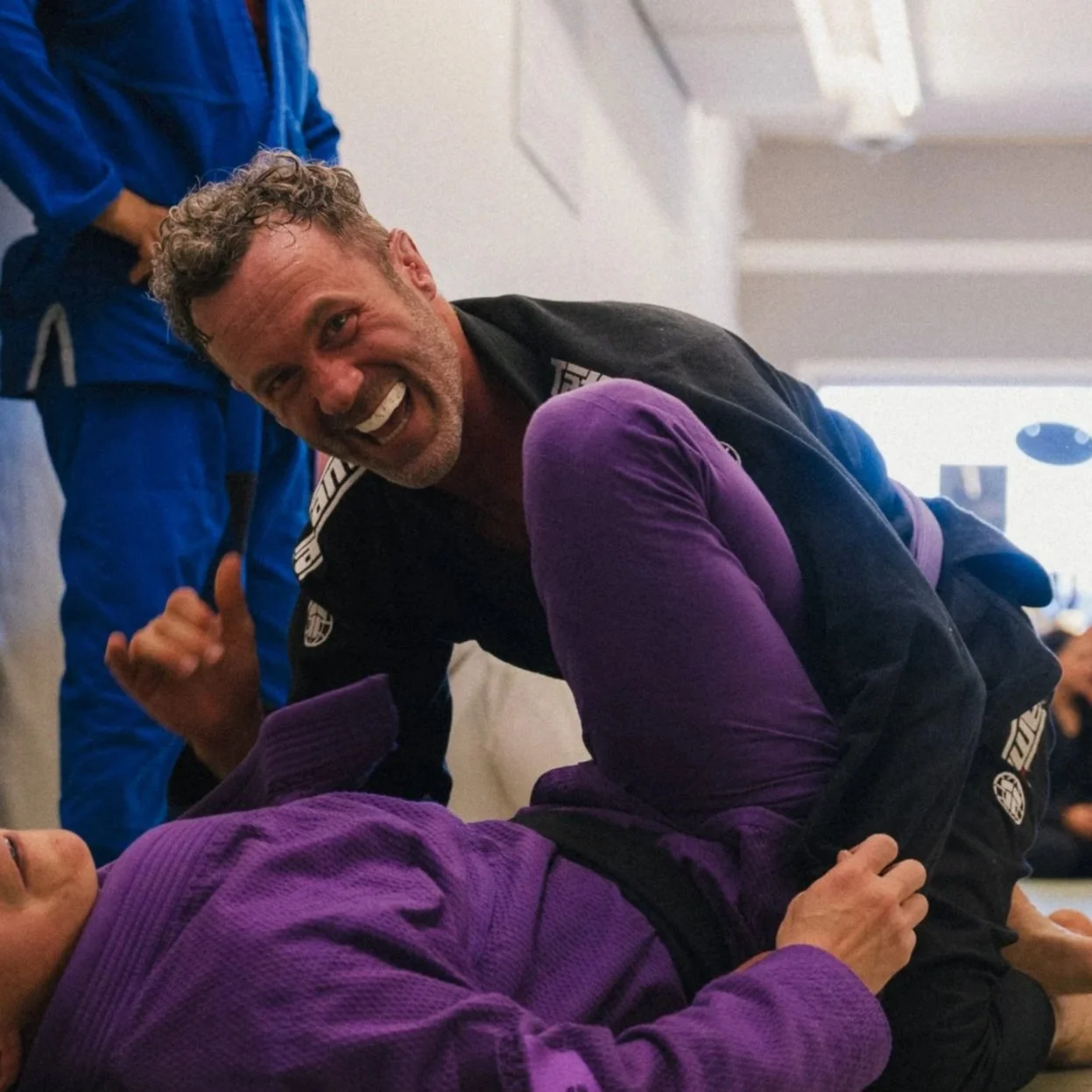 Two people practicing Brazilian Jiu-Jitsu on the mats, with one person in a black gi and the other in a purple gi, smiling and giving a thumbs-up in Avant-Guard Brazillian Jiu Jitsu in Longmont, Colorado.