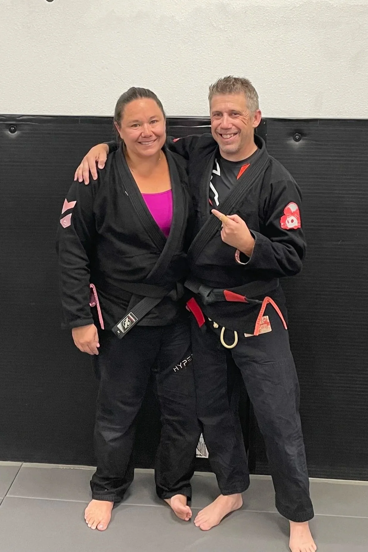 Two people in Brazilian Jiu-Jitsu gis posing together in a martial arts gym, smiling, with one person pointing at the other's gi, both without shoes, against a black padded wall.