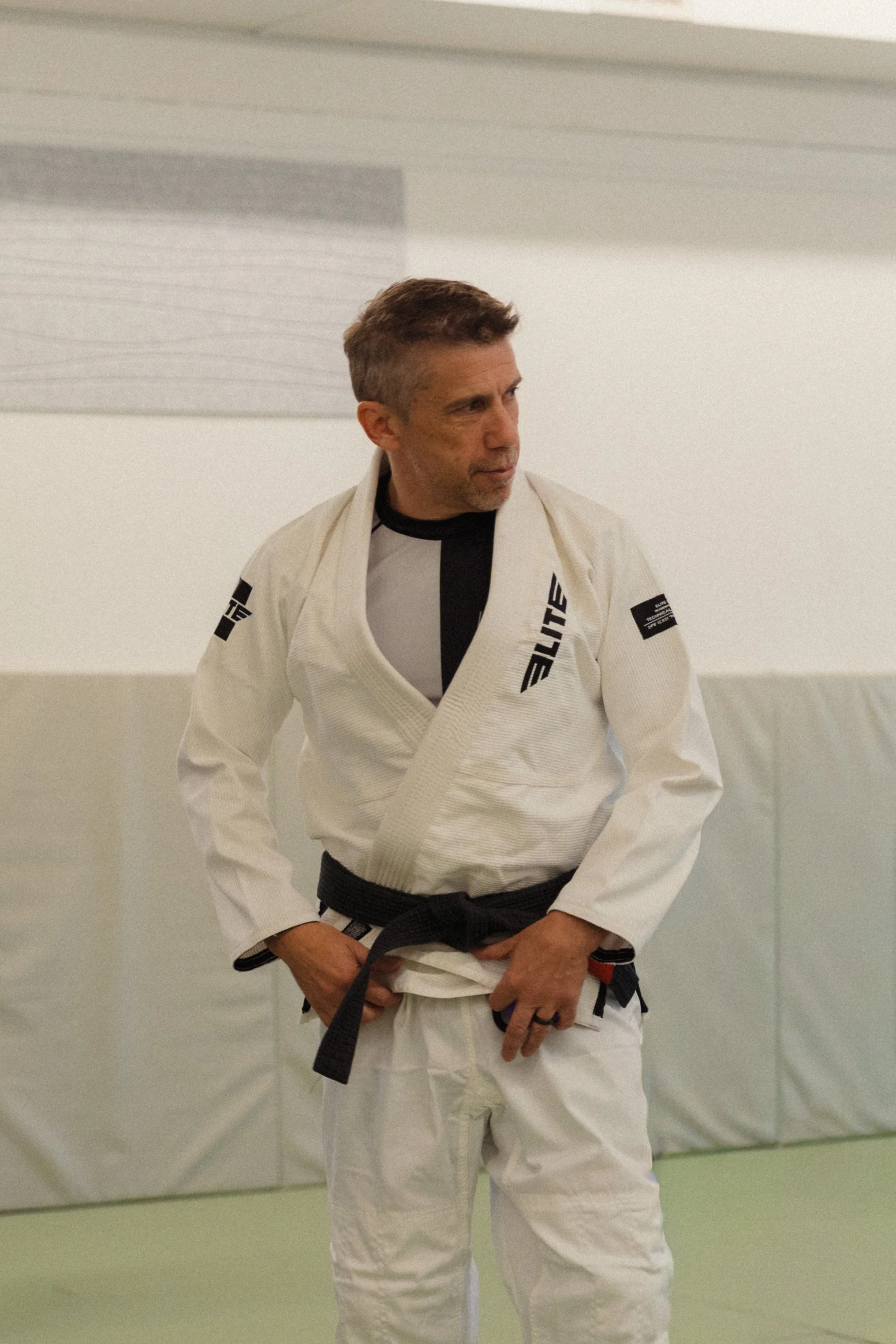A man in a white Brazilian Jiu-Jitsu gi with black belt standing in a training room.