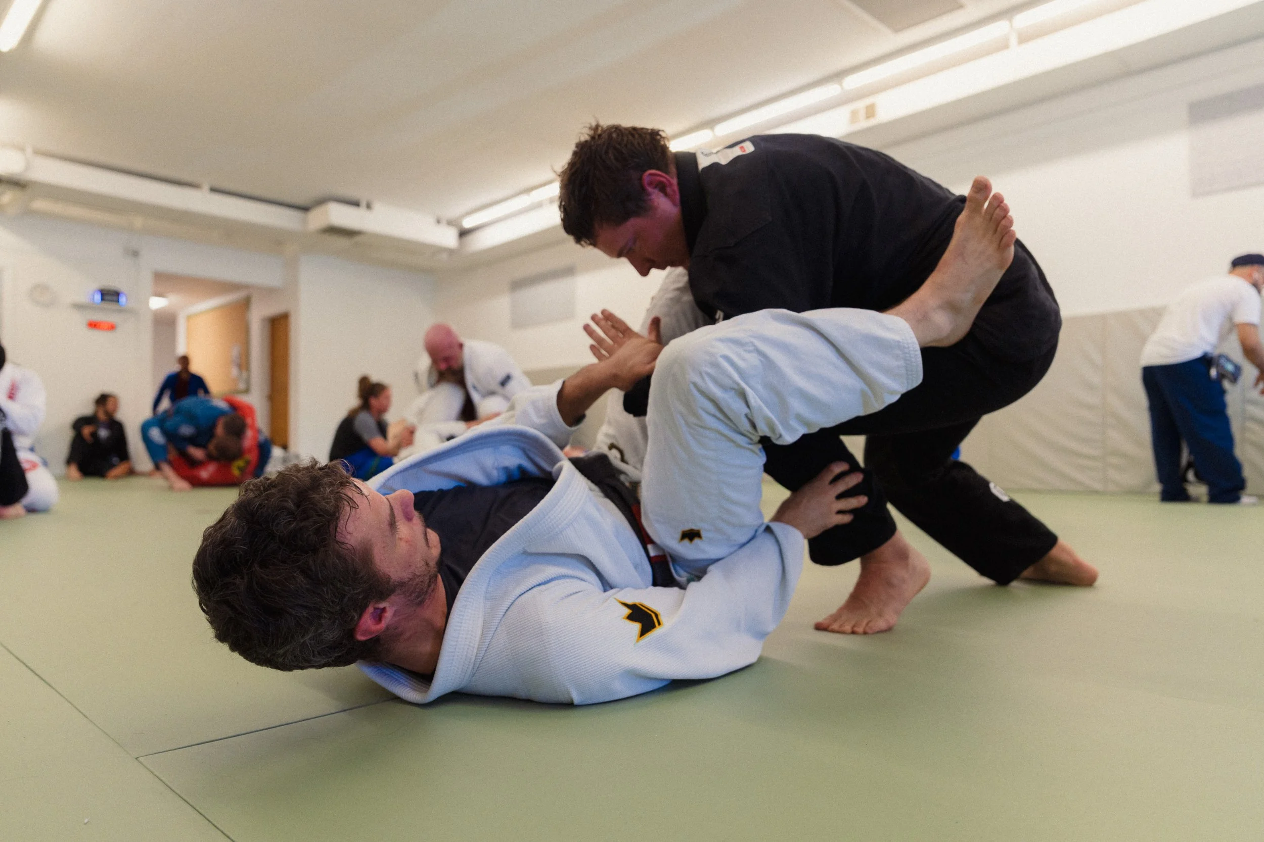 Martial arts practitioners practicing ground fighting techniques in a gym, with one person in a black gi and the other in a white gi on a green mat, with others in the background.