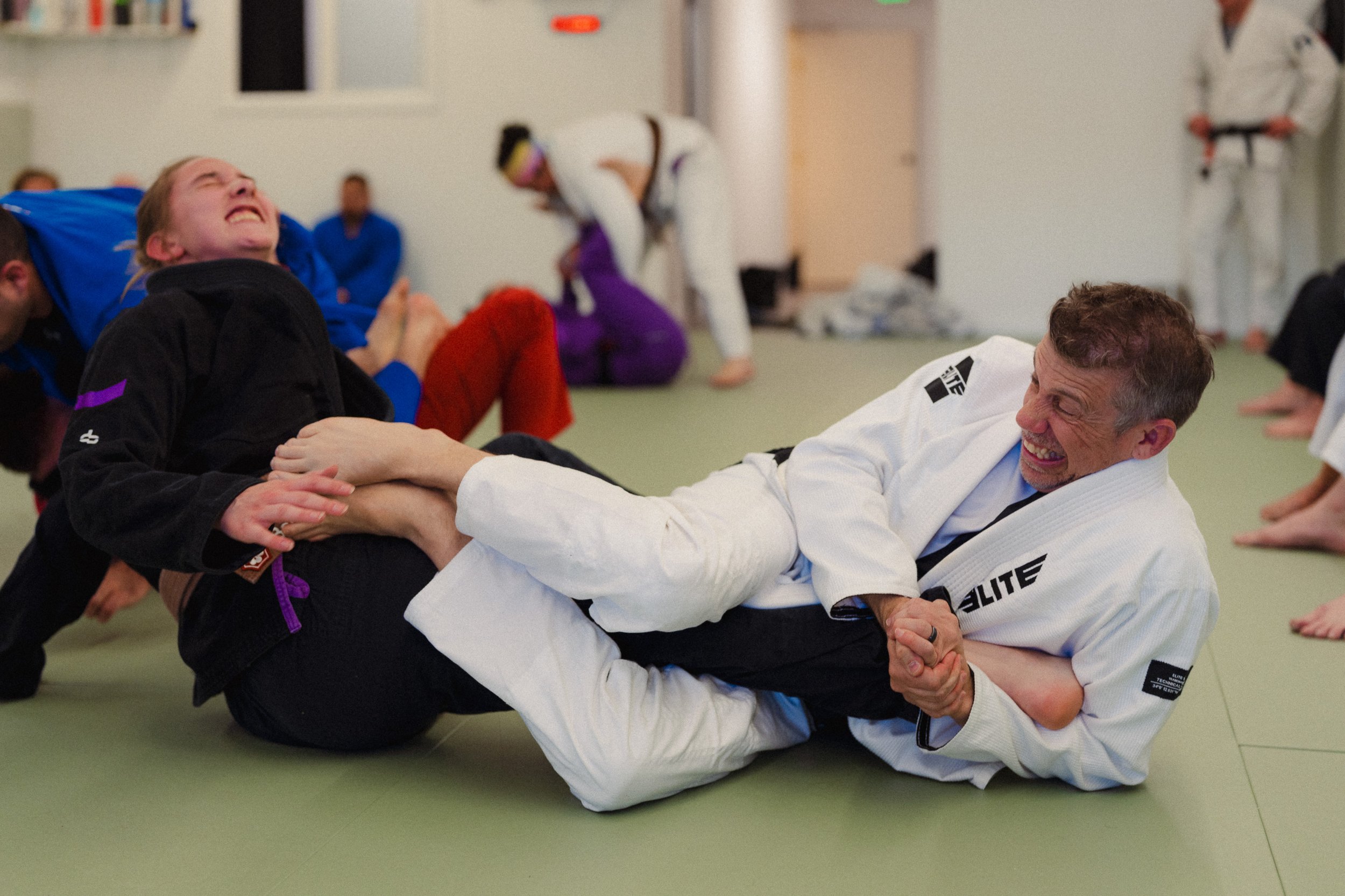 Two martial artists, a man and a woman, practicing Brazilian Jiu-Jitsu on the floor of a martial arts dojo. The man, wearing a white gi, is smiling and gripping the woman, who is wearing a black gi, in a grappling position. Other individuals in gi ar