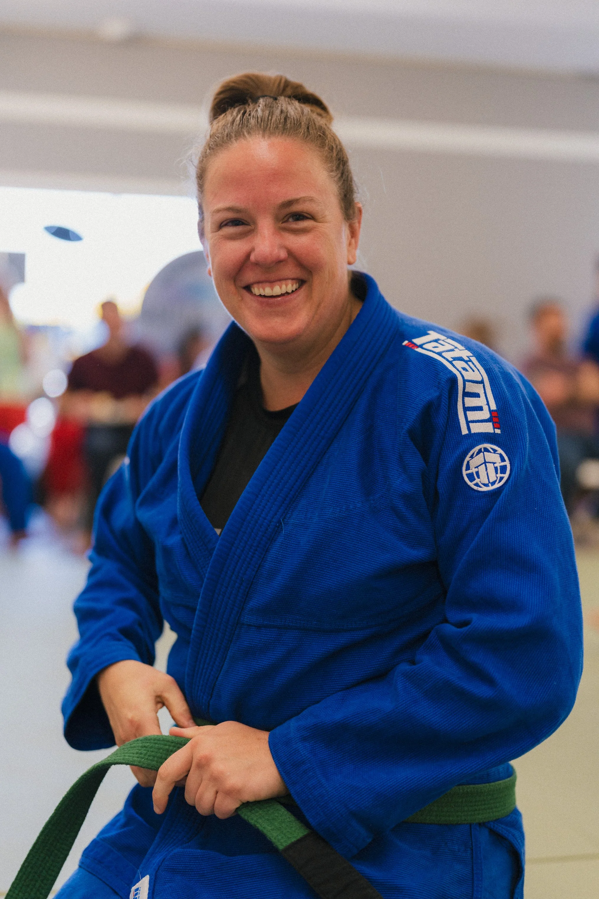 A smiling woman with a bun hairstyle wearing a blue Brazilian Jiu-Jitsu gi, tying her green belt in a martial arts training center with blurred people in the background.