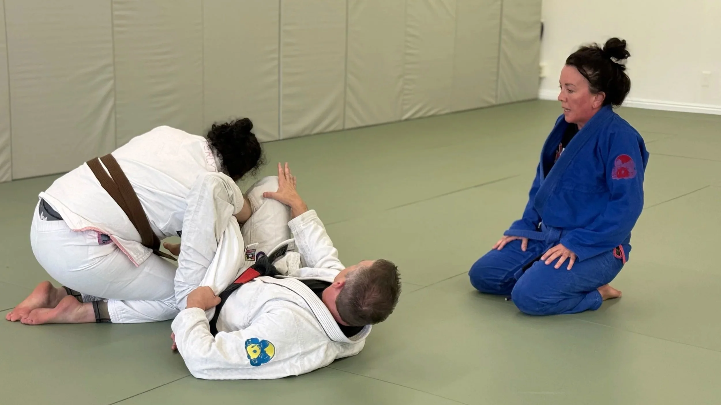 Two women practicing Brazilian Jiu-Jitsu on a padded mat, with one woman wearing a blue gi kneeling and the other wearing a white gi on top of a man, demonstrating a grappling technique.