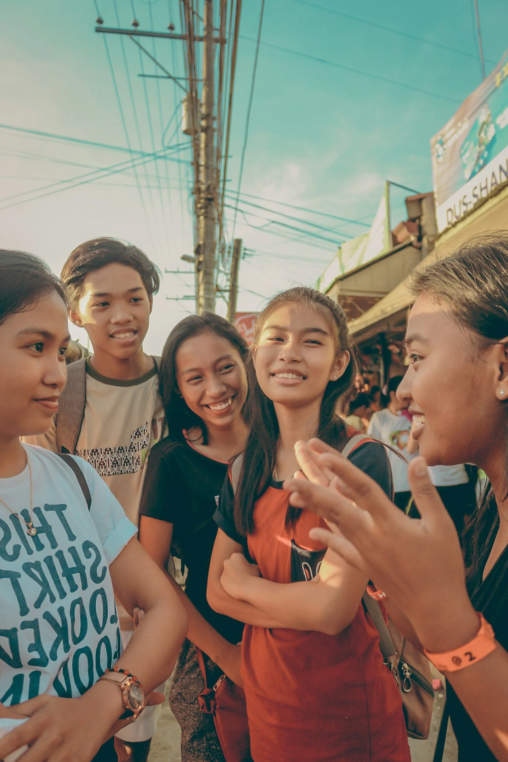 Group of young people gathered outdoors, smiling and conversing with each other. There are power lines and a signboard in the background.