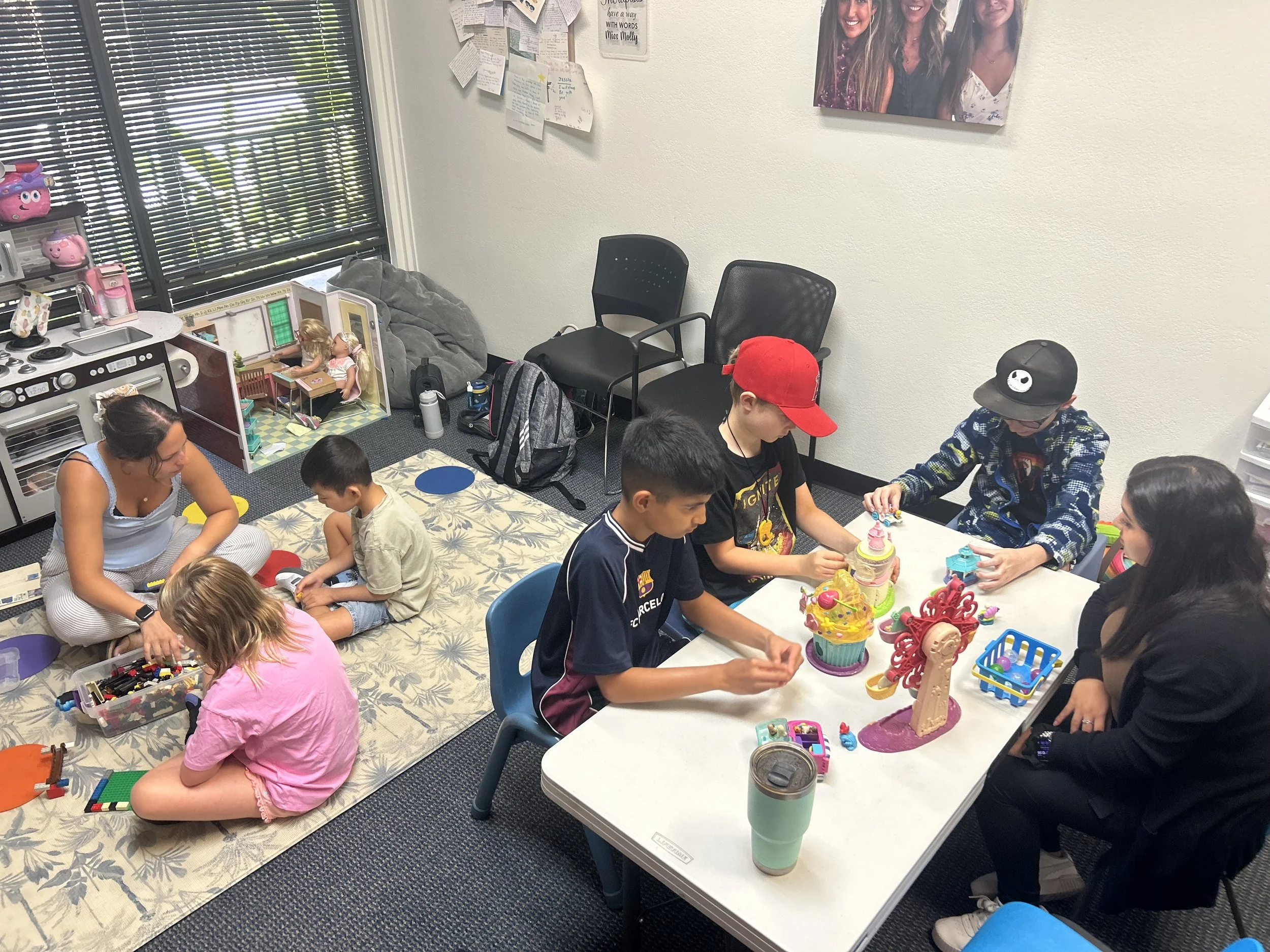 Children and adults playing with toys and LEGO bricks in a room decorated with photos and notes on the wall, with a large window with blinds letting in natural light.