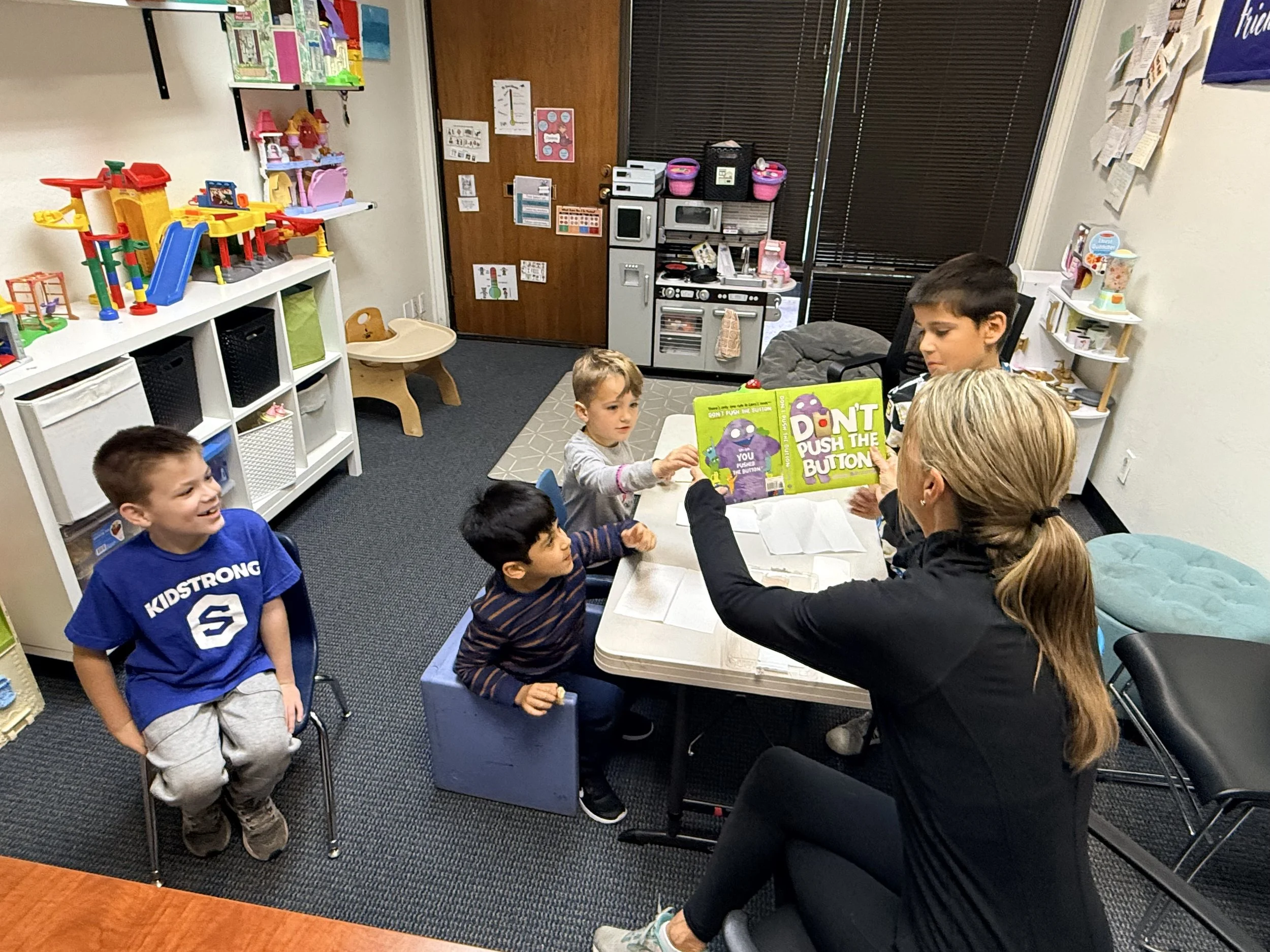 A group of children and adults are gathered in a classroom or playroom, engaging in an activity with a children's book titled 'Don't Push the Button!' The children are sitting or standing around a table, and there are toys, shelves, and posters on the walls, with a small toy kitchen in the background.