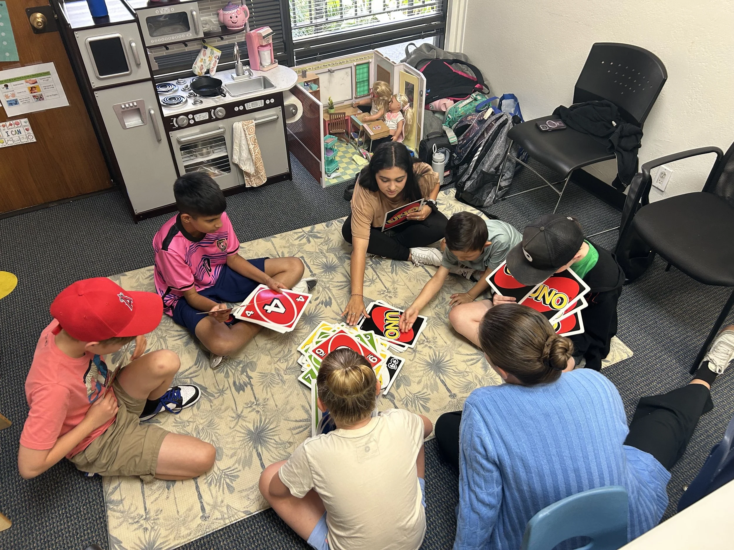 A group of children sitting on a carpeted floor playing Uno cards while a woman helps them. The room resembles a small play kitchen with a toy stove, sink, and small kitchen accessories, and backpacks and chairs are against the wall.