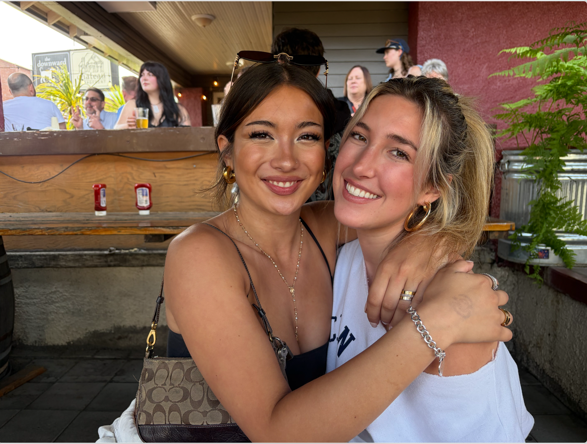 Two young women hugging and smiling at the camera in an outdoor patio setting. They are socializing outside.