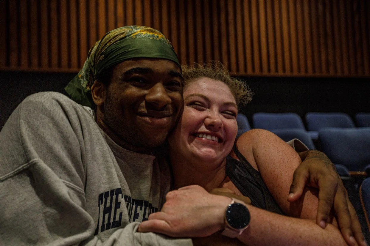Two people sitting close together, smiling happily in an auditorium with blue seats and wooden paneling.