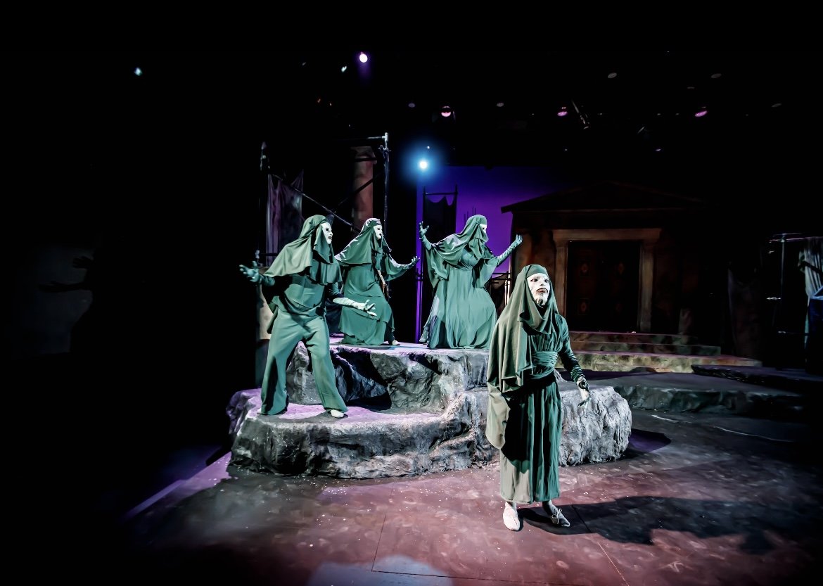 Theatrical production of 'The Taming of the Shrew' with actors dressed as nuns on stage, with some gesturing and one in the foreground looking upward, illuminated by stage lights.