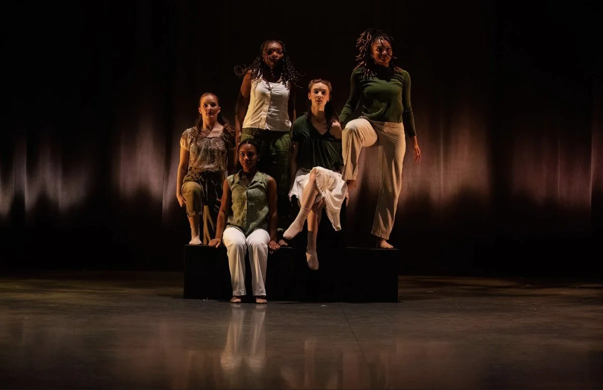 Six women dressed in casual clothing on a dark stage, striking poses with some standing, sitting, and one balancing on a box.