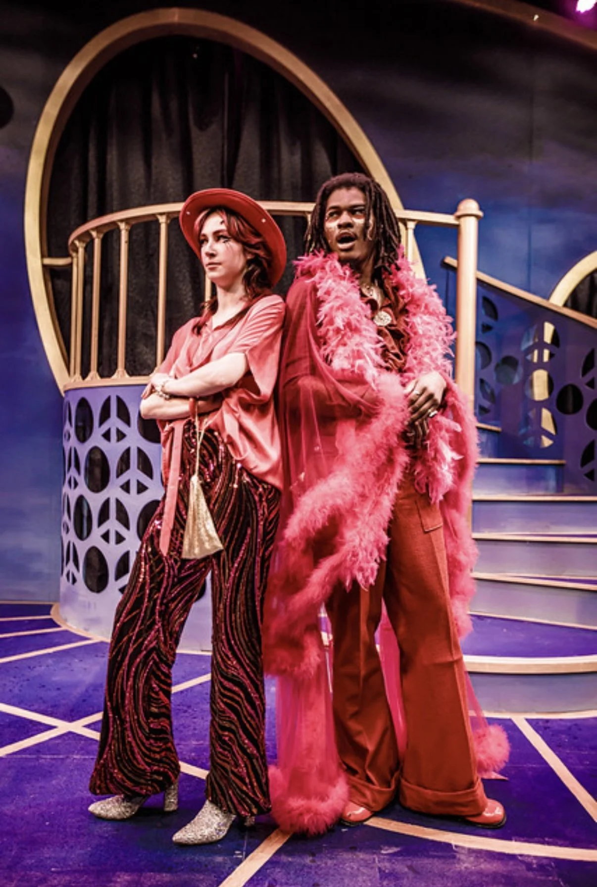 Two people dressed in retro, vibrant clothing stand in front of a whimsical, colorful set with purple, pink, and gold accents and a black curtain backdrop.