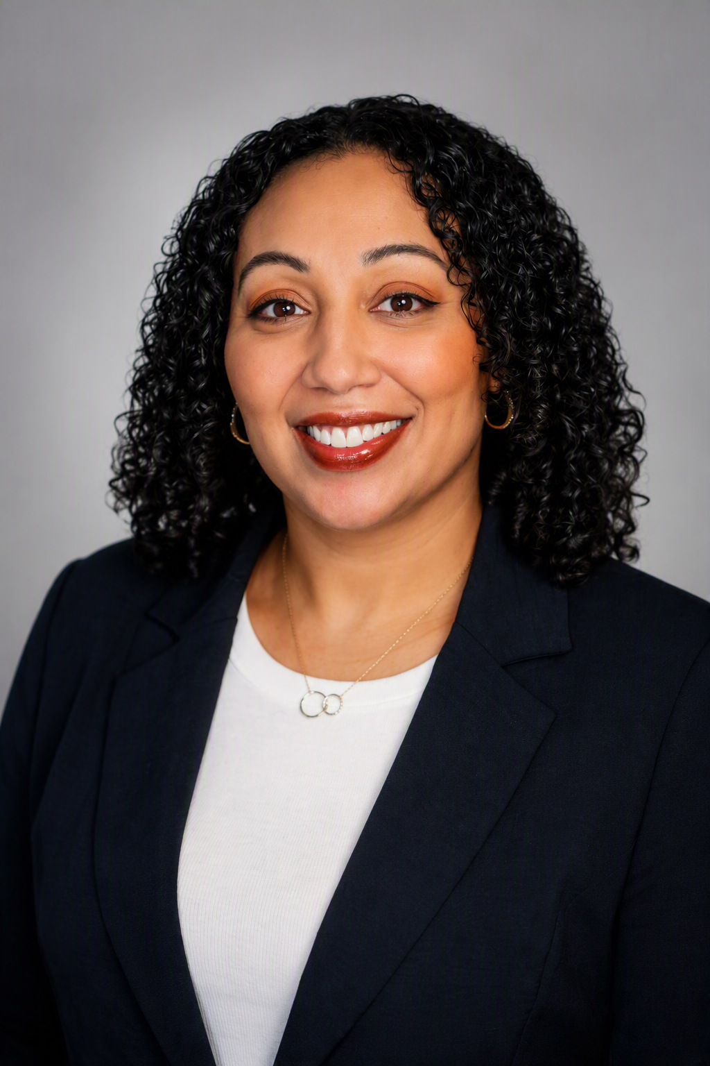A professional woman with curly black hair smiling, wearing a black blazer, white top, gold hoop earrings, and a gold necklace, against a plain grey background.