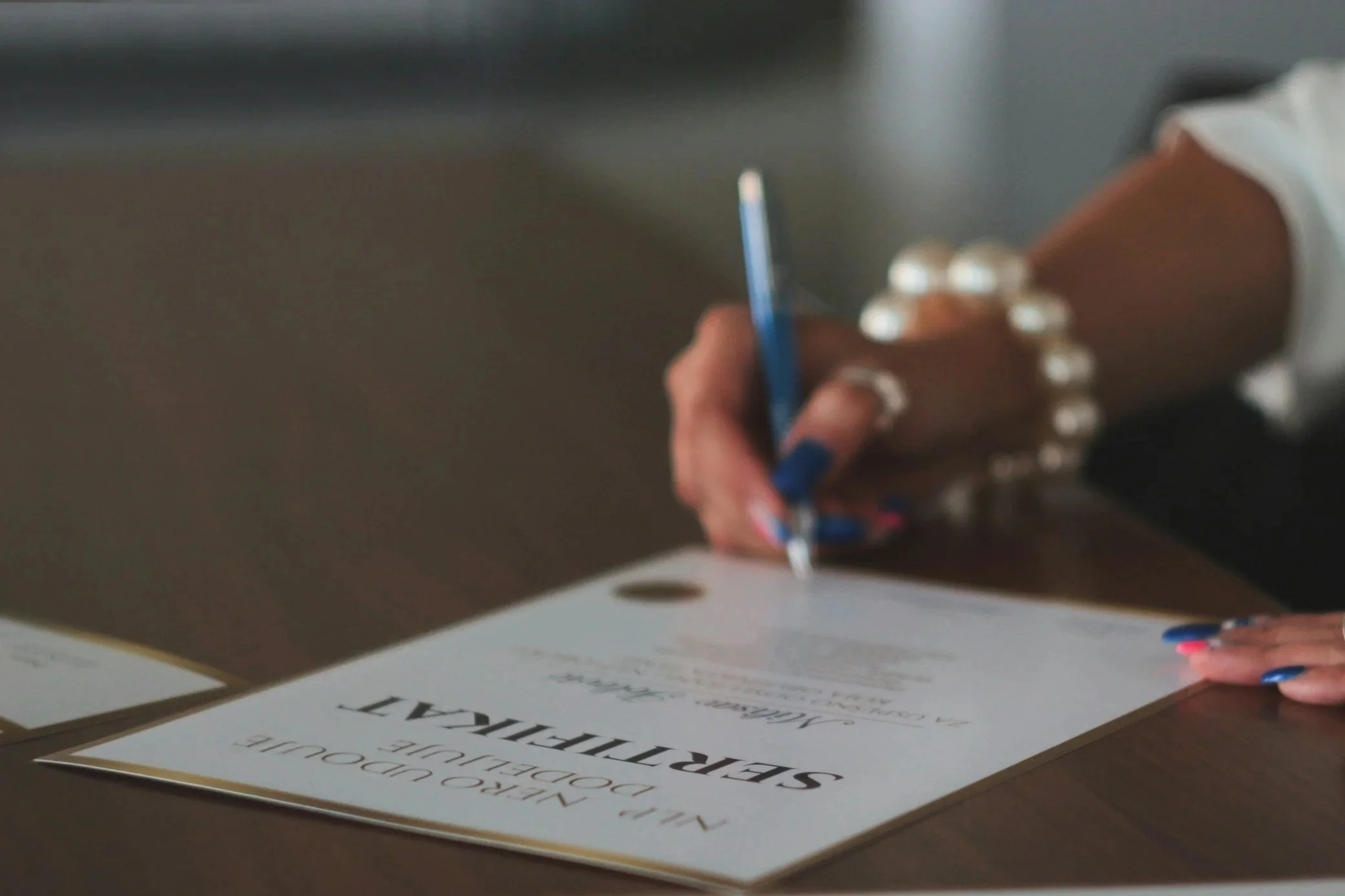 A person wearing a white shirt with a pearl bracelet is signing a document on a table with a blue pen.