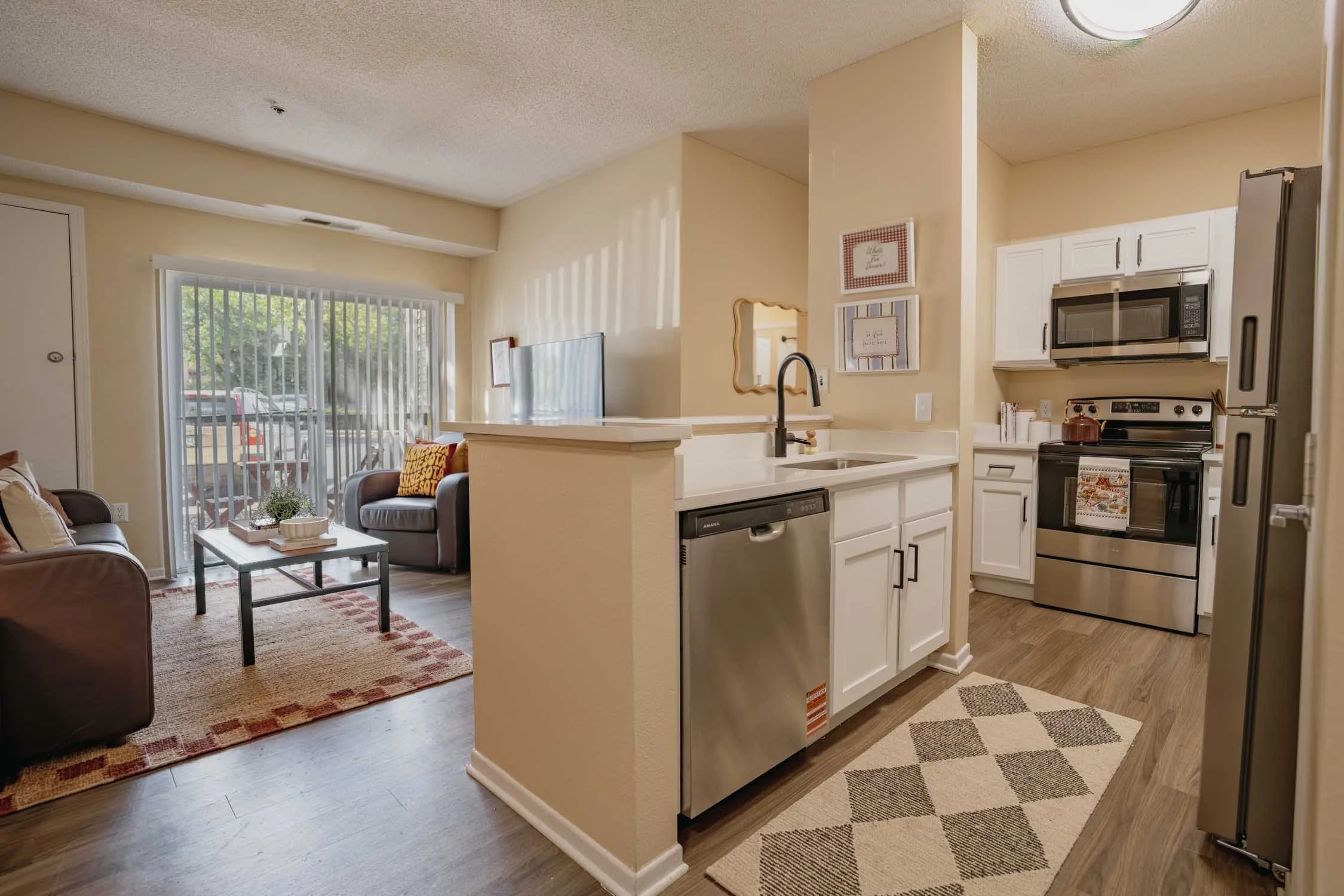 Open-concept living room and kitchen with sliding glass door, beige walls, white cabinetry, stainless steel appliances, and a small beige and white rug.