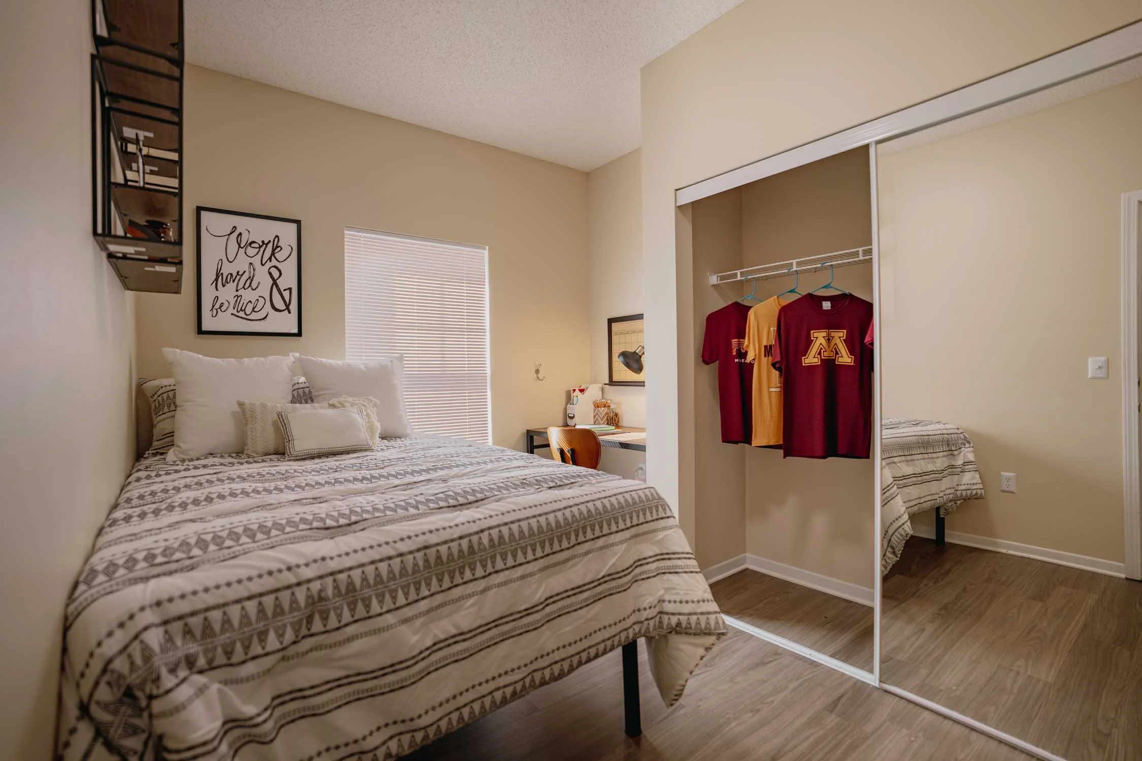 A bedroom with a bed covered in a patterned beige and white bedspread, pillows, a window with blinds, wall art with motivational quote, and an open closet with hanging shirts and a mirror.
