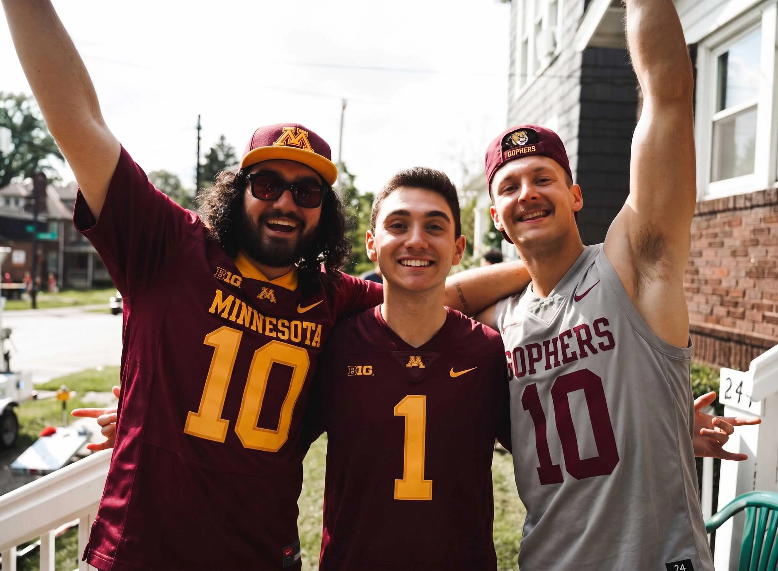 Three young men wearing University of Minnesota and University of South Carolina sports jerseys and hats, smiling and celebrating outdoors.