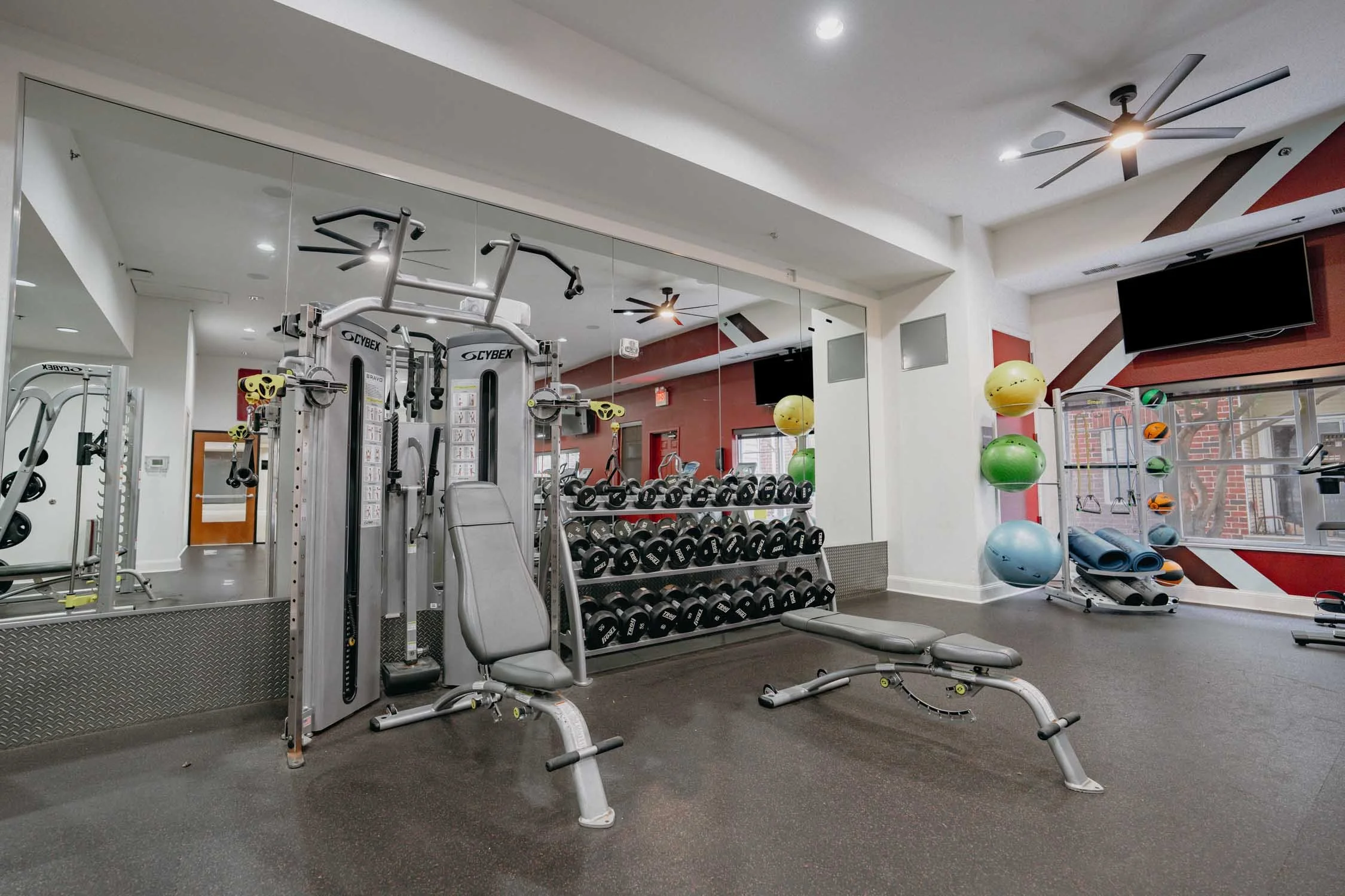 Modern gym with Cybex strength-training equipment, dumbbell racks, exercise balls, benches, mirrors, and wall-mounted TVs