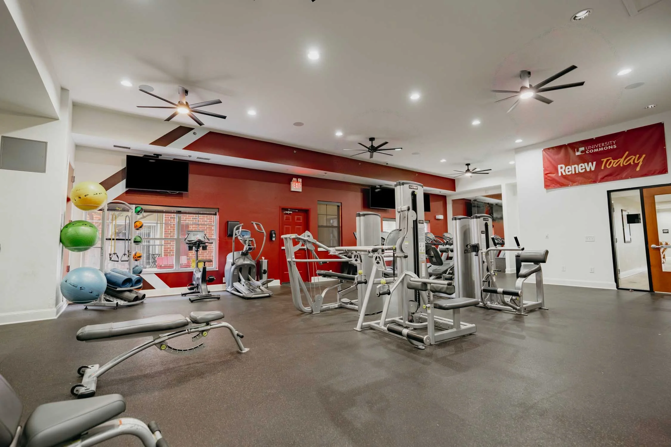 Fitness center with exercise equipment, stability balls, treadmills, and weight machines, with a red wall and ceiling fans.