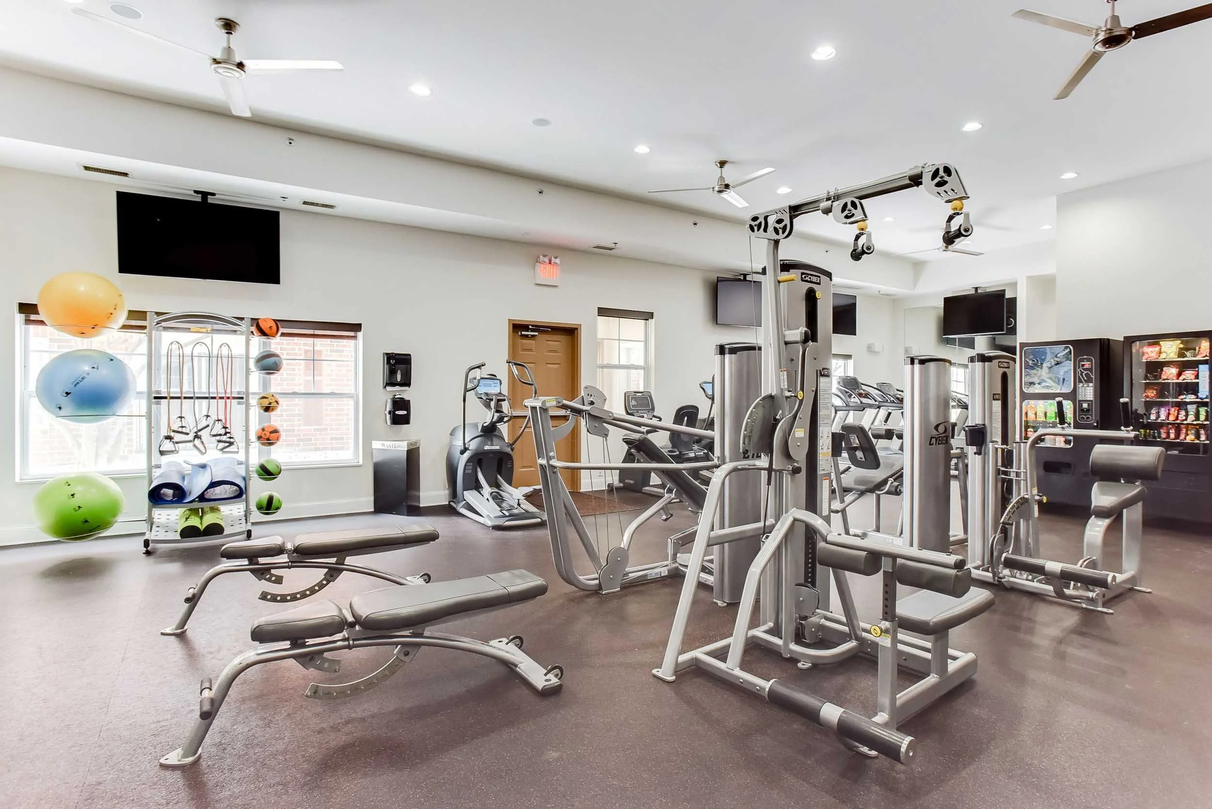 This image shows the interior of a gym with various exercise equipment including cardio machines, weight machines, exercise balls, and a vending machine. The room has white walls, windows, and ceiling fans.