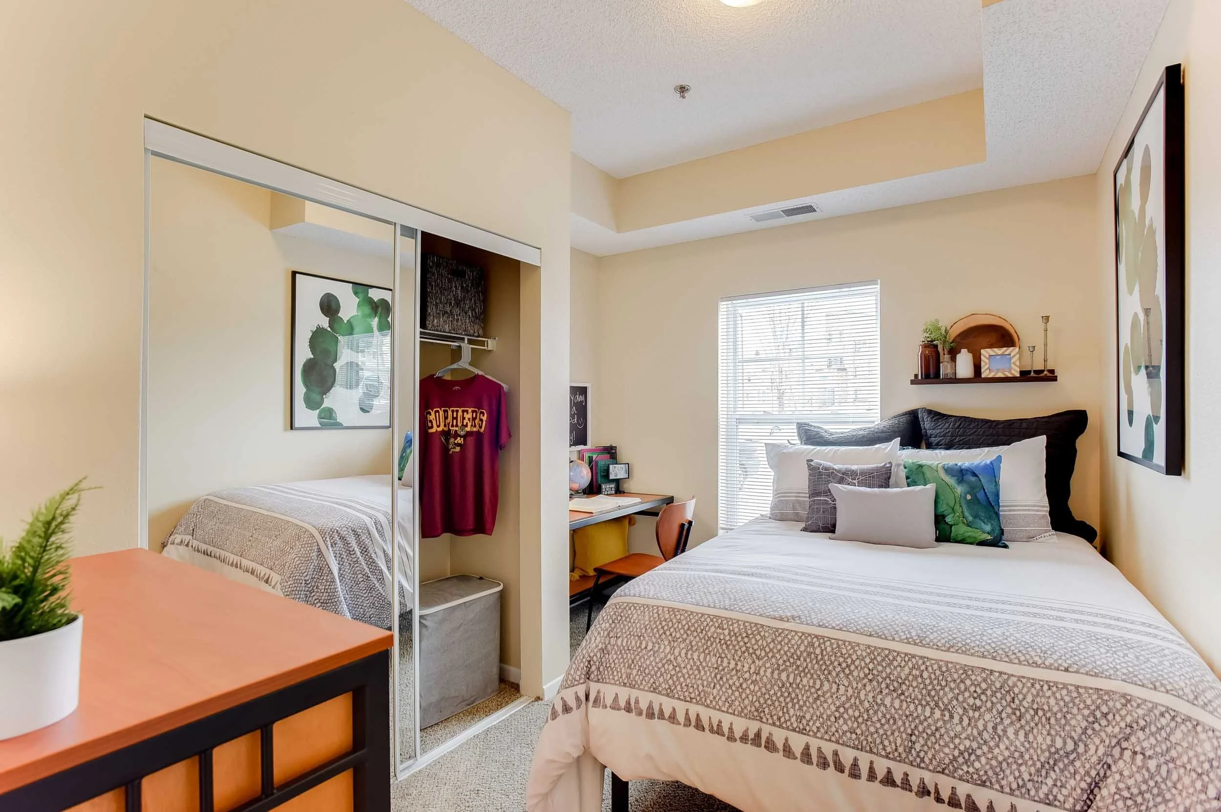 A cozy bedroom with two beds, a small desk with a chair, a window with blinds, and decorative items on a wall shelf.