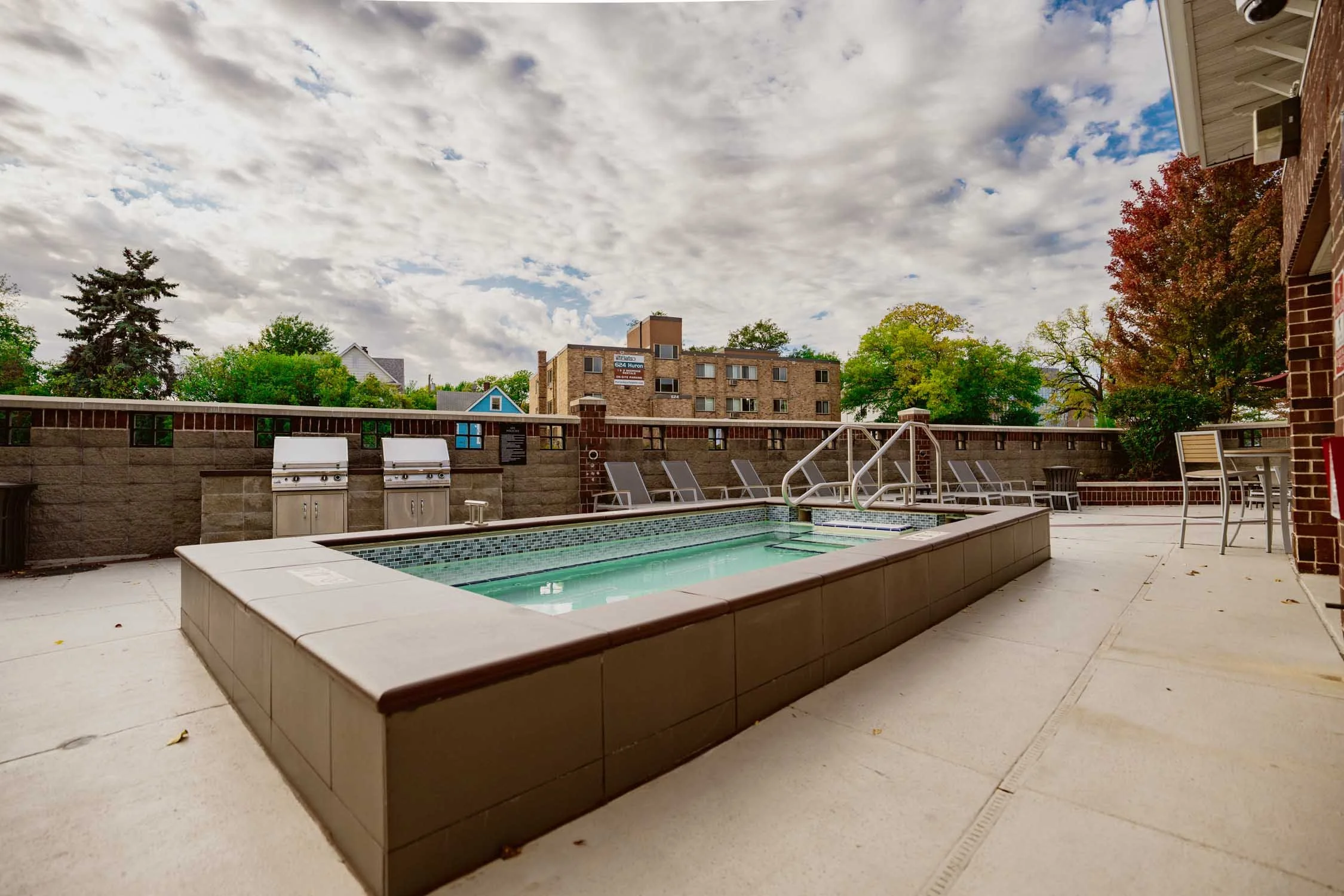 Oversized hot tub area with lounge chairs, a brick wall, trees with fall foliage, and a cloudy sky.