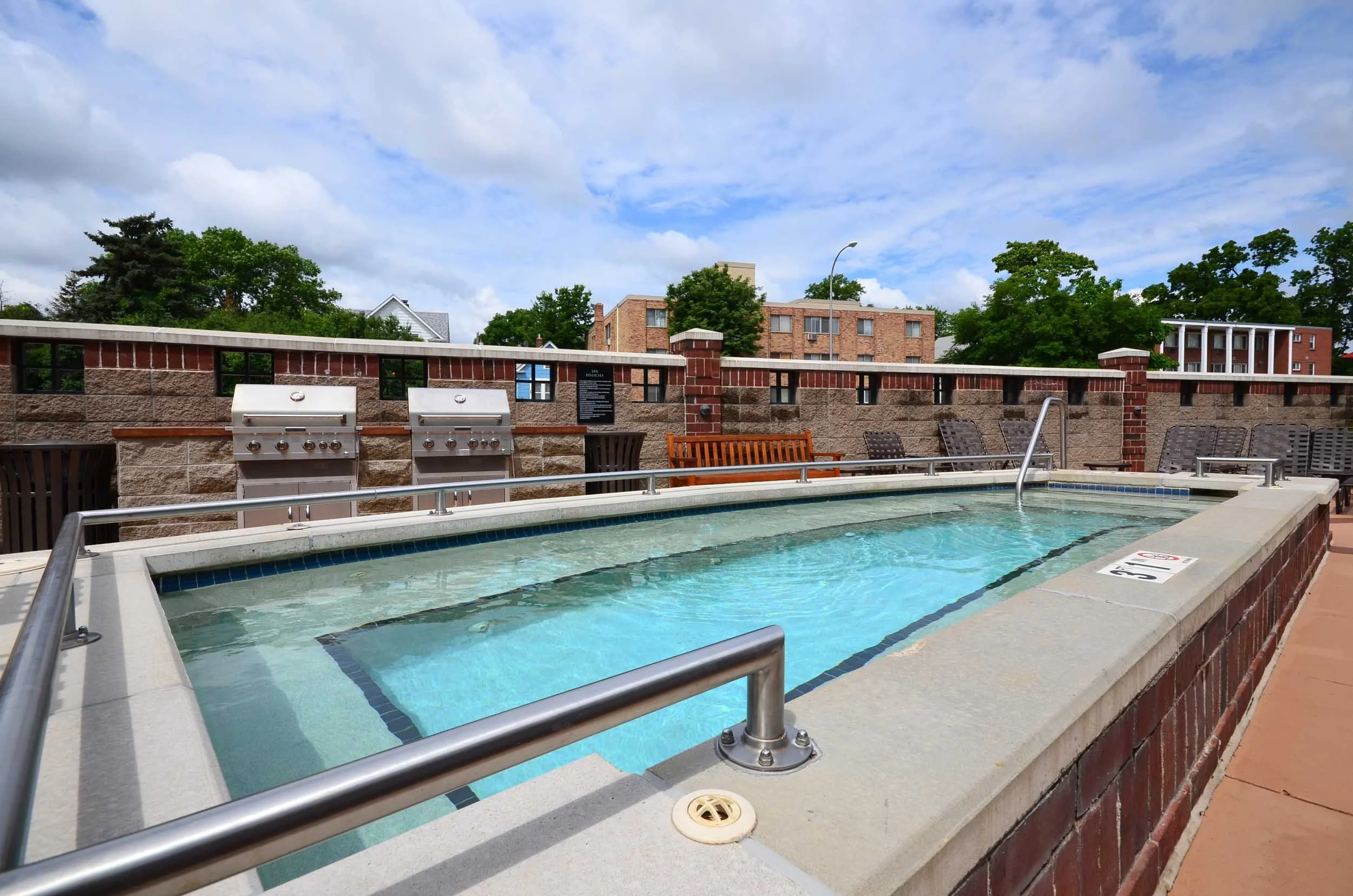 View of a rooftop hot tub with a brick wall and city buildings and trees in the background under a partly cloudy sky.